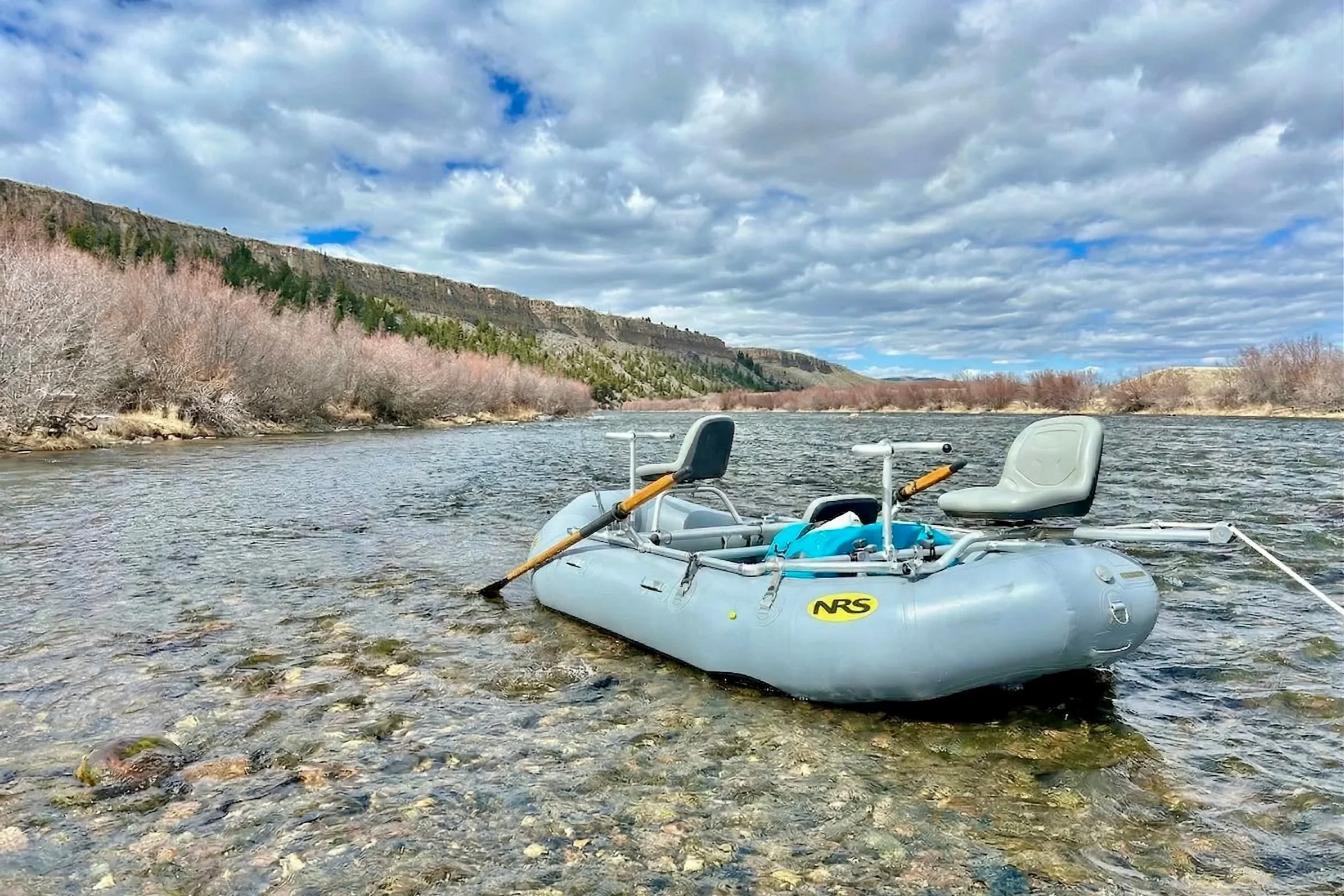 An inflatable raft with two seats floating on a shallow river with a rocky bed, surrounded by leafless trees and a hillside with green vegetation and cliffs under a partly cloudy sky.