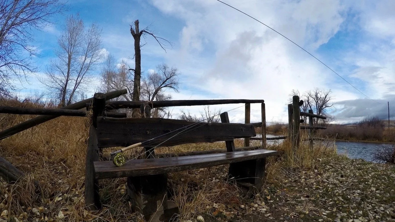 A fly rod resting on a wooden bench at Williams Bridge overlooking the Gallatin River, representing the relaxed and professional guided fly fishing trips offered by Flyshot Outfitters in Bozeman, MT.