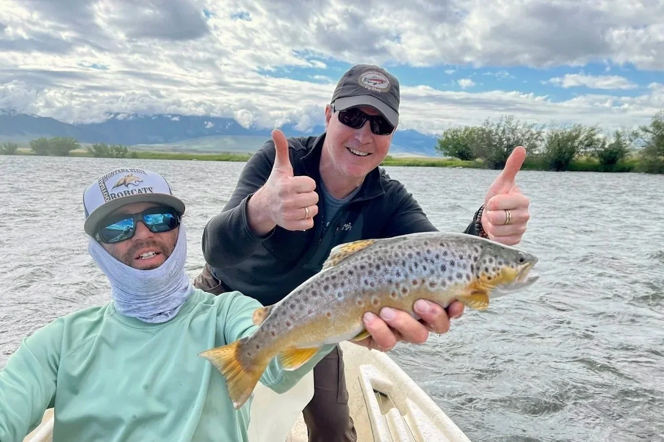 Two men on a boat holding a large spotted fish, with one man giving a thumbs-up and the other smiling with a fishing hat and sunglasses, lake and mountains in the background.