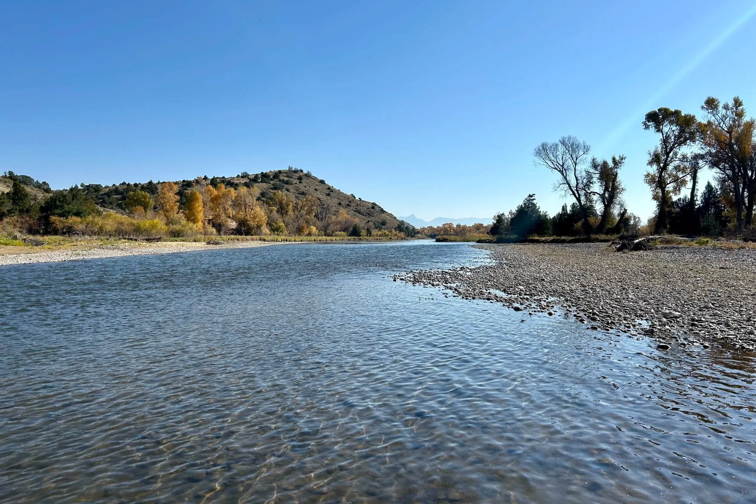 A river flows through a landscape with trees showing fall foliage, a hillside on the left, and a bright blue sky overhead.