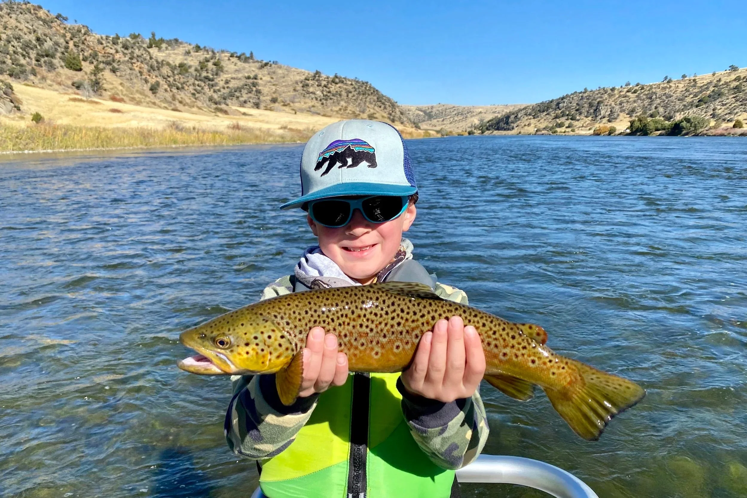 A smiling young angler wearing a life jacket and polarized sunglasses holding a large brown trout during a safe, family-friendly half-day guided float trip on the Lower Madison River near Bozeman.