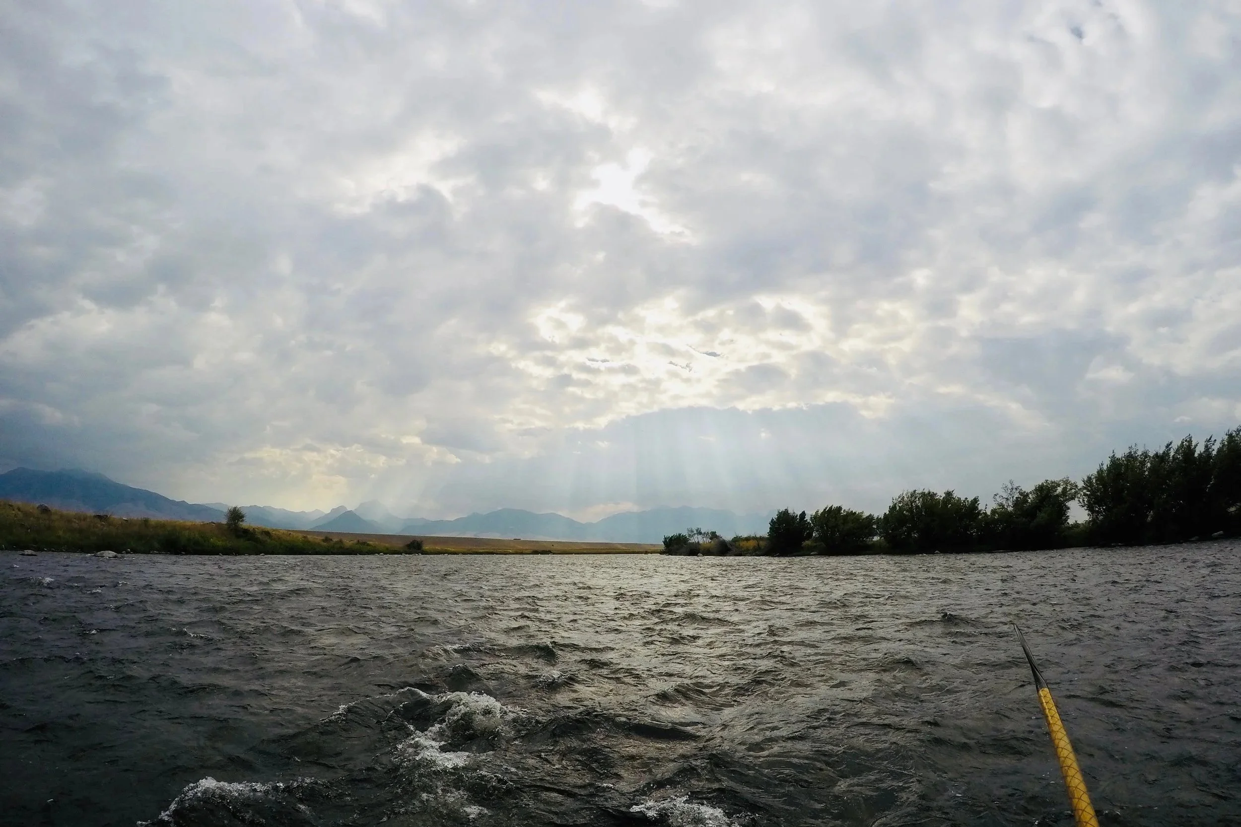 Dramatic sun rays breaking through overcast storm clouds on the Madison River, featuring a handcrafted Sawyer oar in the foreground during a professional drift boat trip in Montana.
