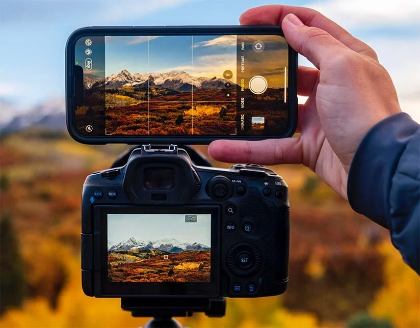 A person is taking a photograph of a mountain landscape with a DSLR camera and a smartphone, both capturing a scene of mountains, trees, and an orange sunset sky.