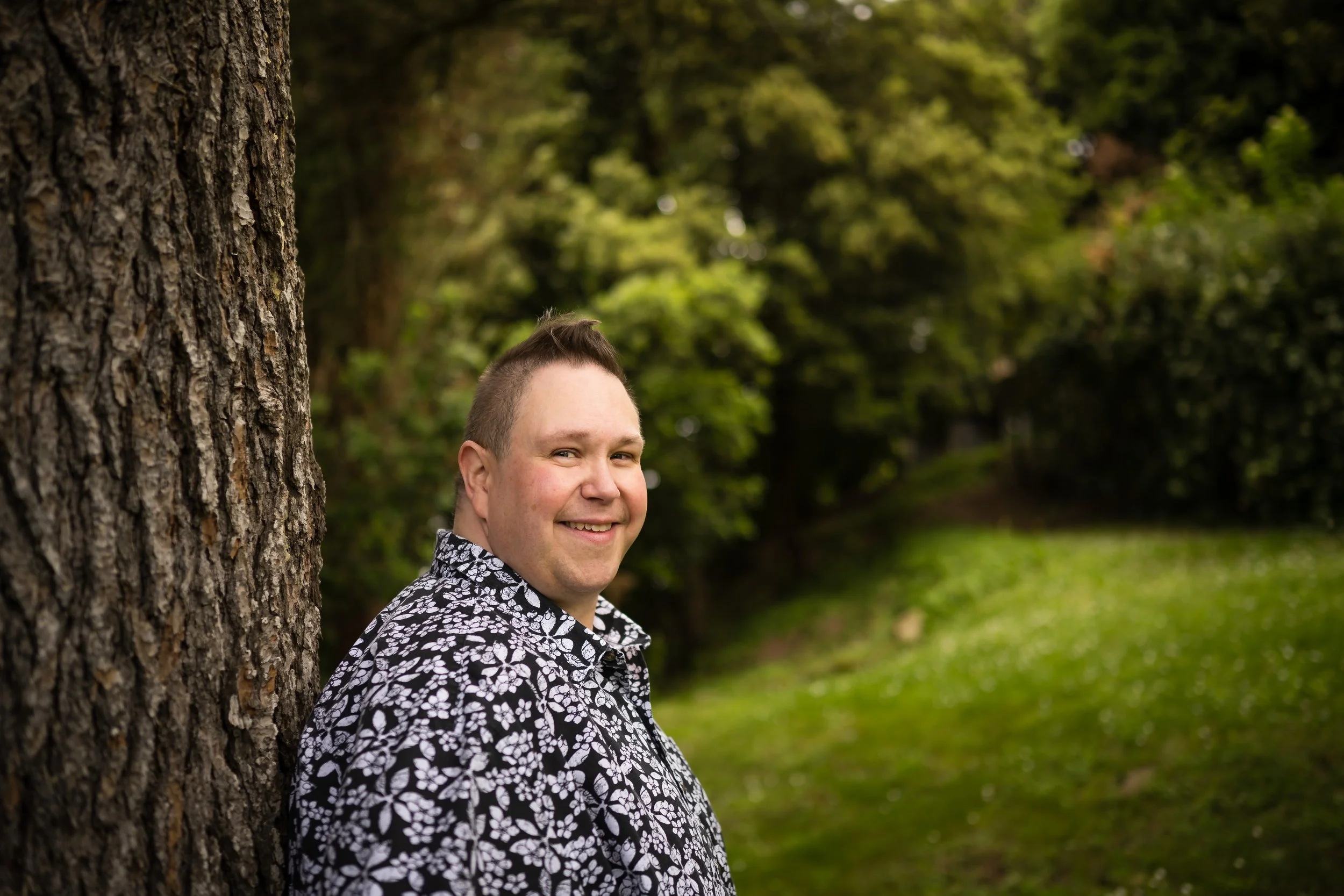 James Allen Hall leaning against a tree, wearing a black and white floral shirt.