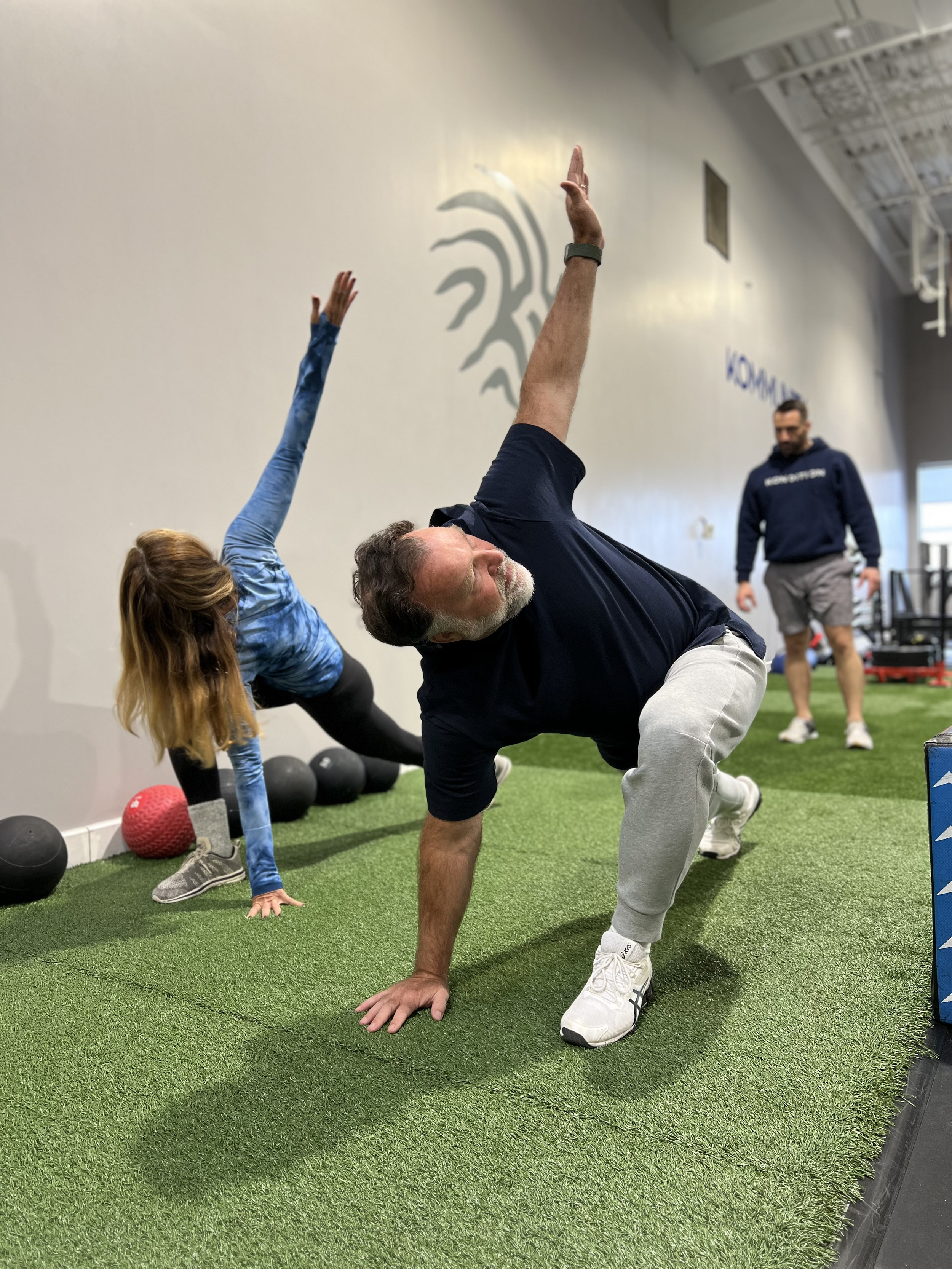 Man in black t-shirt and gray sweatpants doing a yoga pose on a green turf, with a woman in blue workout clothes and man in hoodie in background, inside a gym.