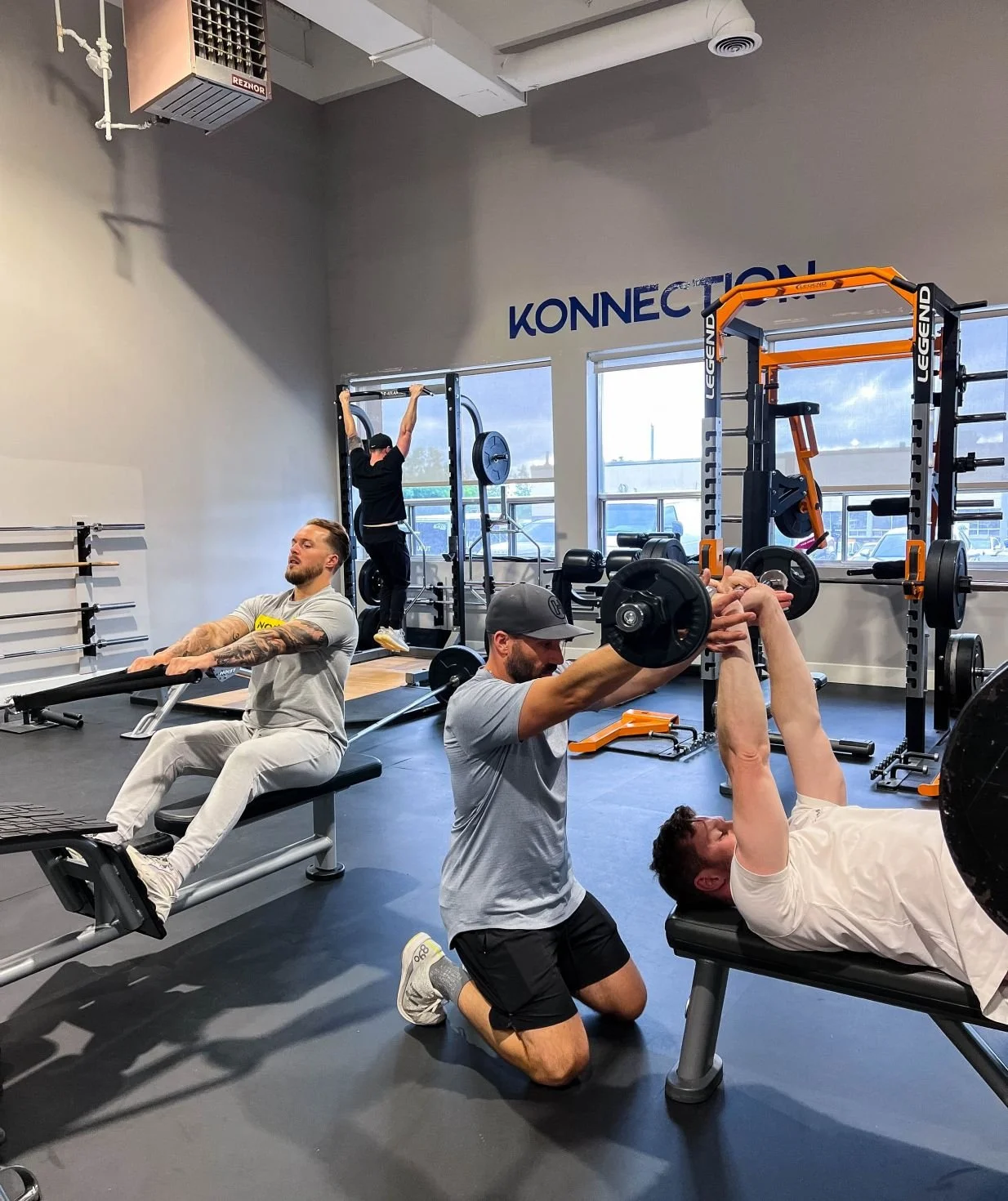 Three men working out in a gym, with one lying on a bench pressing a barbell, another kneeling and lifting a dumbbell, and a third in the background doing pull-ups. There are gym equipment and windows in the background.