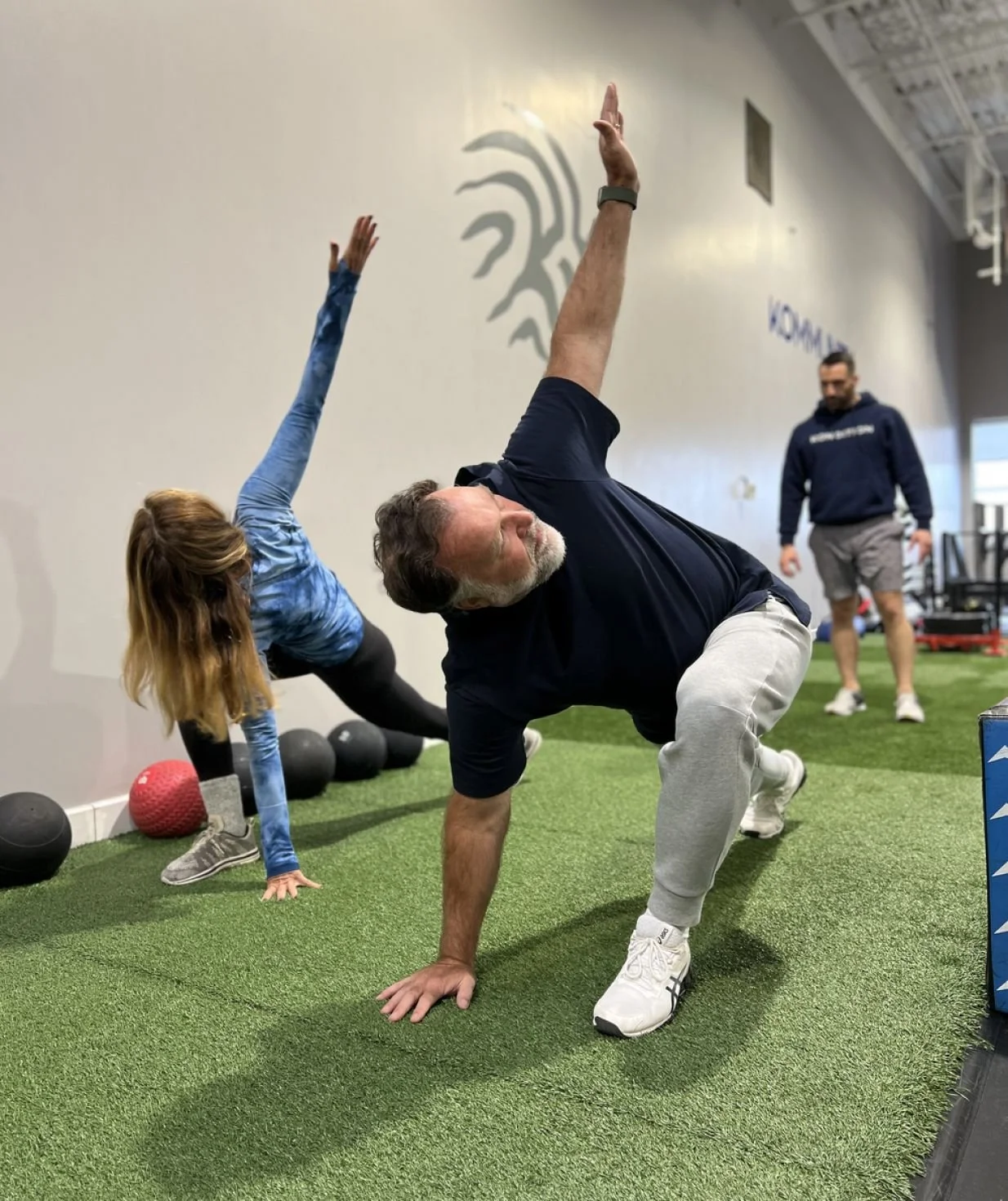 Two people exercising on artificial turf with a trainer overseeing, one man in a bear crawl position and a woman reaching towards the ceiling in a stretch, with weights lined along the wall.