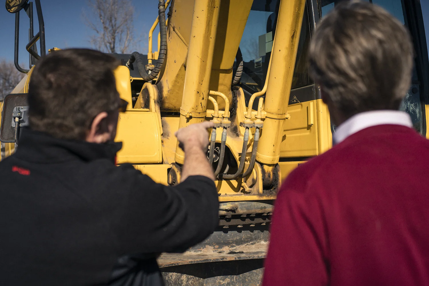 Anneal Business Coaching graphic from Martin Holland showing a worker pointing to the hydraulic lines on a yellow excavator while a coworker looks on.