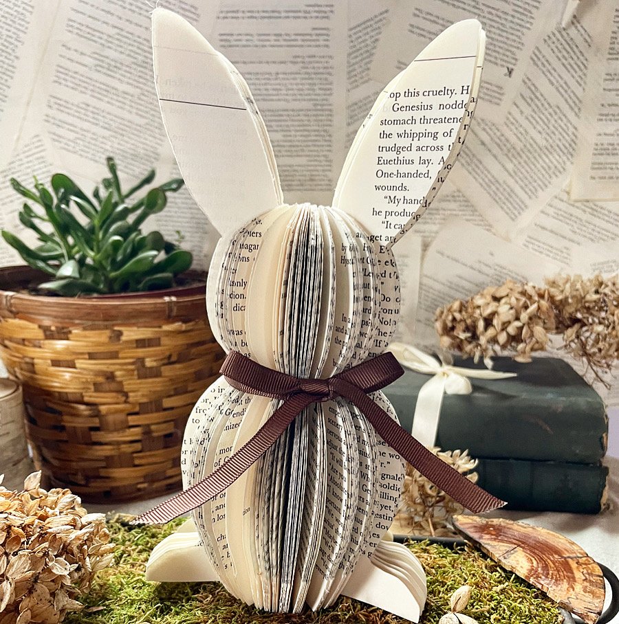 An upcycled book rabbit sculpture sits on a bed of moss with a backdrop of plants, books and book pages