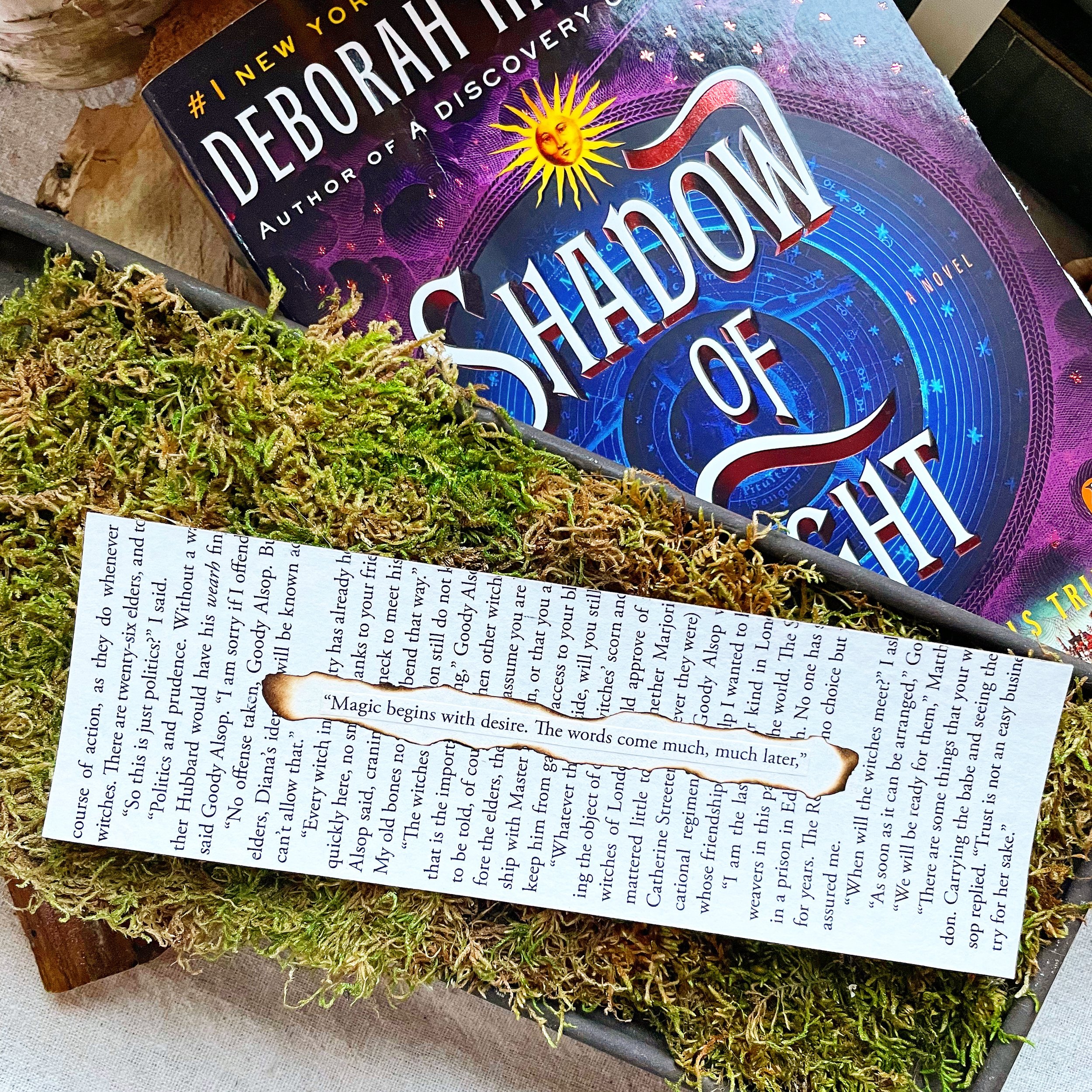 A bookmark made of upcycled book pages with the quote: "Magic begins with desire. The words come much, much later." With the book Shadow of Night behind it