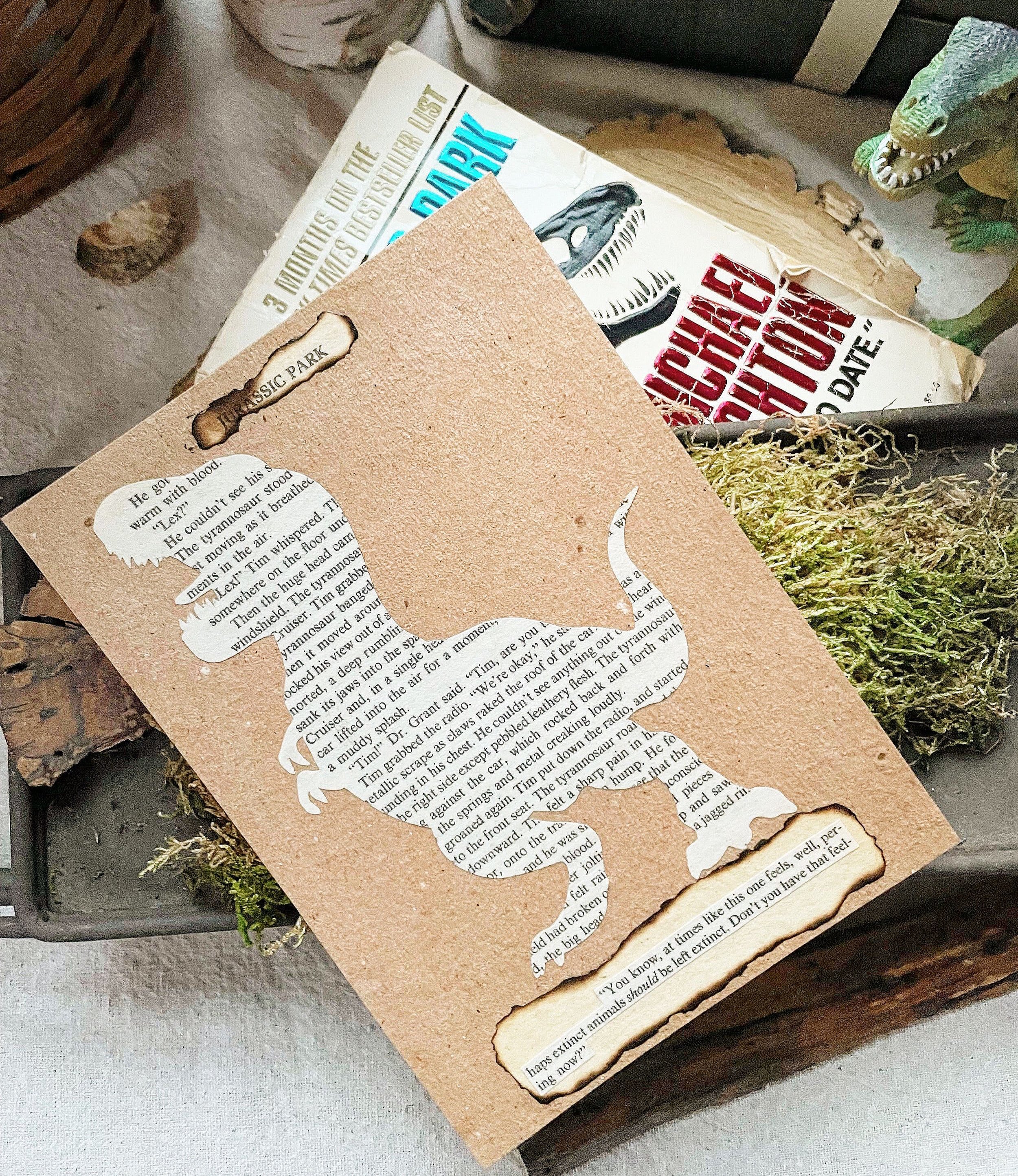 A print featuring a silhouette of a t-rex cut from book pages along with a quote and the title of jurassic Park sits on a bed of moss with the Jurassic Park novel behind it