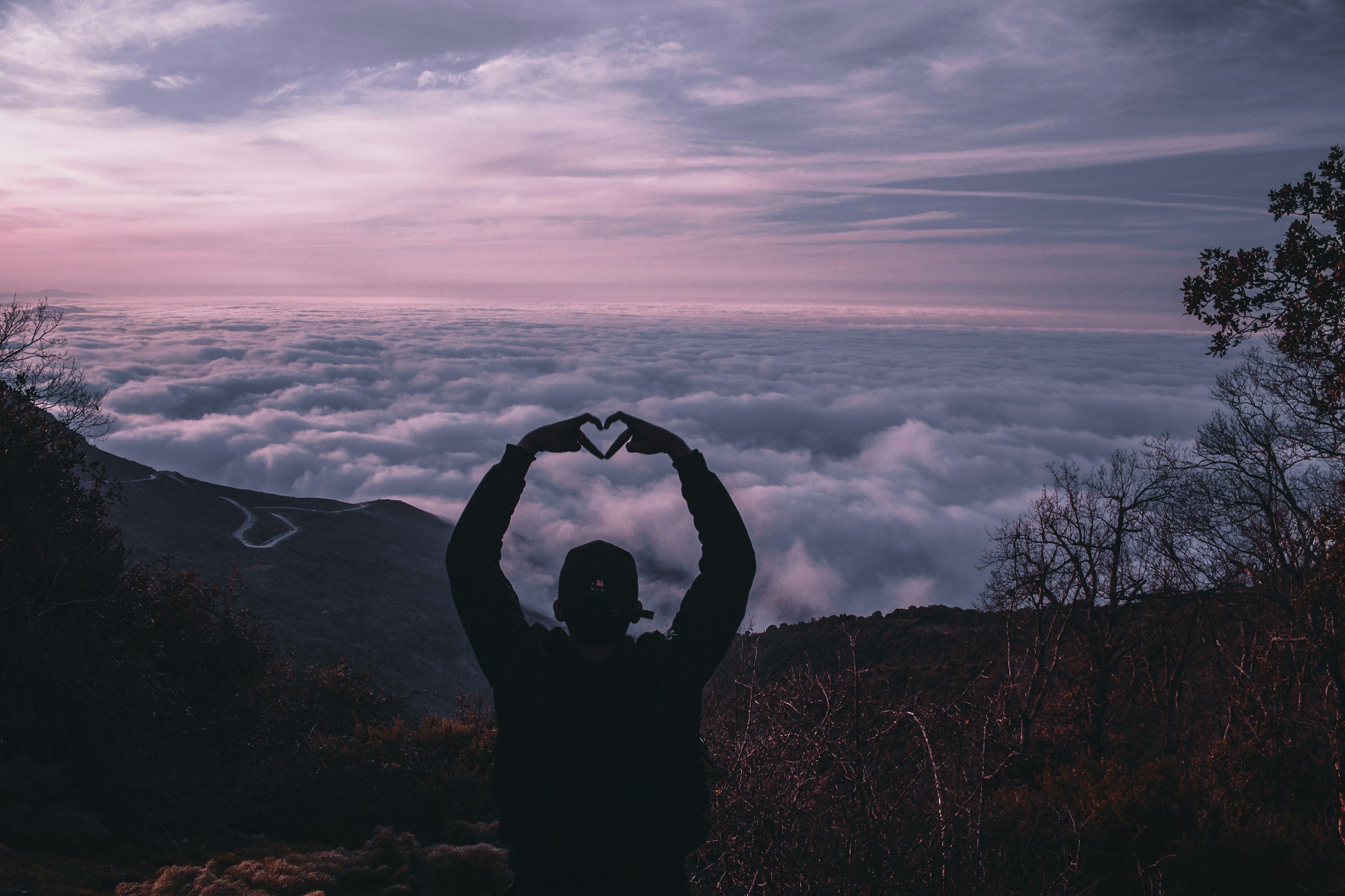 Silhouette of a person making a heart shape with their hands, standing on a hill above clouds at sunrise or sunset with pink, purple, and blue sky.