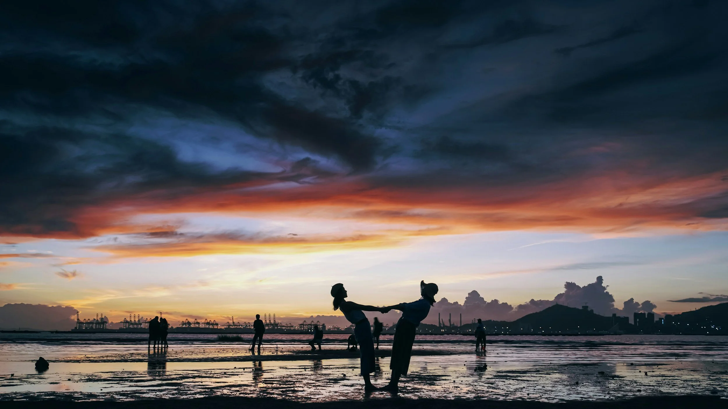 Silhouettes of children playing on the beach at sunset with a colorful sky, distant people, and industrial structures in the background.