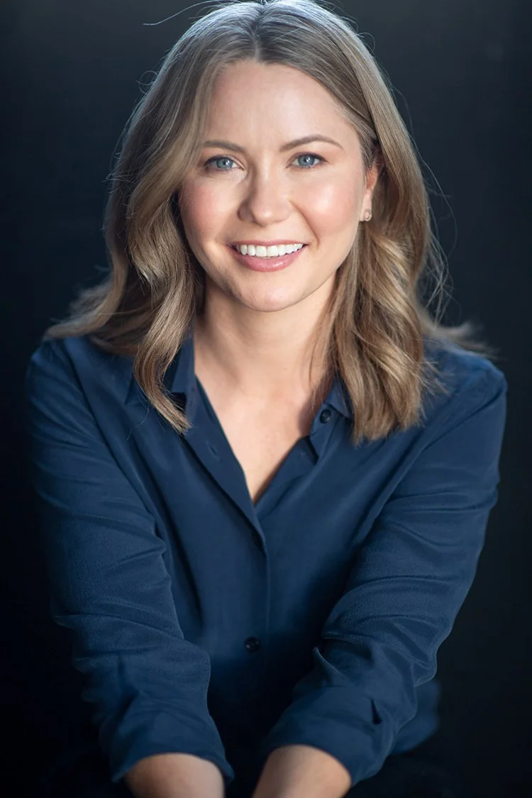 A young woman with shoulder-length blonde hair and blue eyes, smiling and wearing a dark blue button-up shirt, against a dark background.