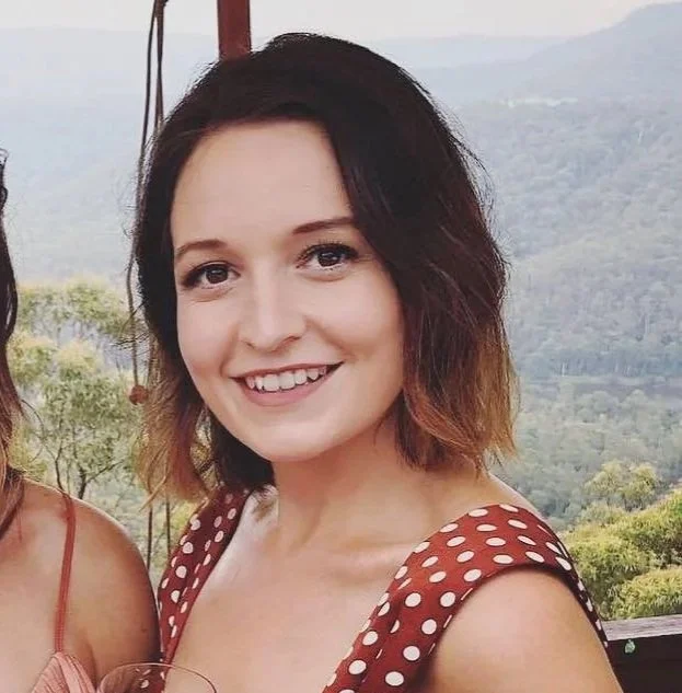 A woman with shoulder-length hair smiling outdoors with scenic mountains and greenery in the background.