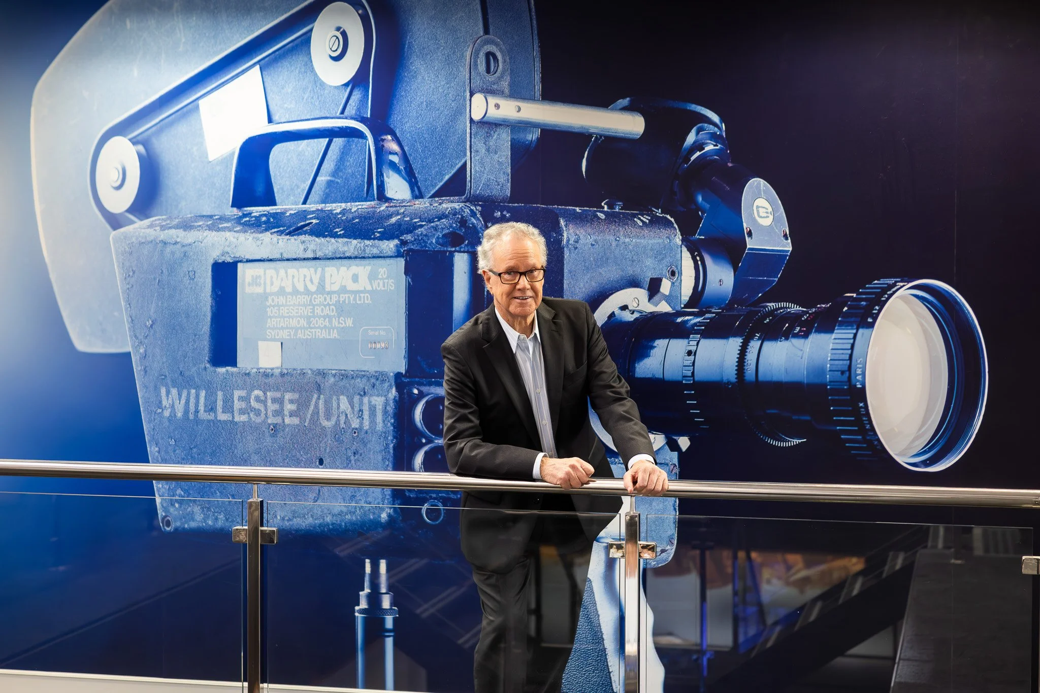 Terry Willesee stands in front of a mural featuring the historic Willesee Unit camera at Channel 9 Perth, symbolising his long-standing contribution to broadcast journalism — photographed by Ryan Ammon | Ammon Creative.