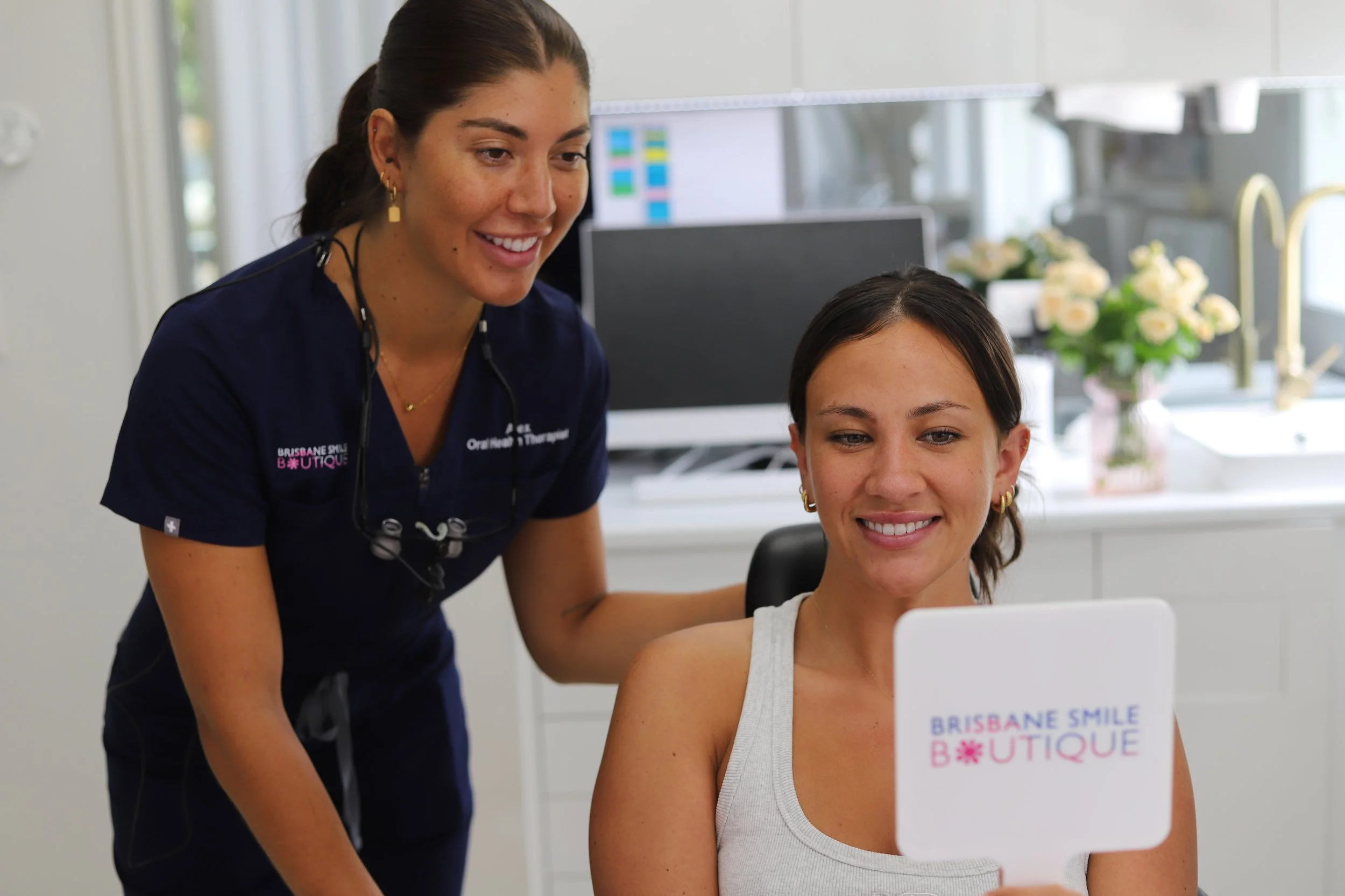 Female with brunette hair and white smile in scrubs with name tag that says Brisbane Smile Boutique and Oral health therapist talking to a patient who is looking in a mirror that says Brisbane Smile Boutique
