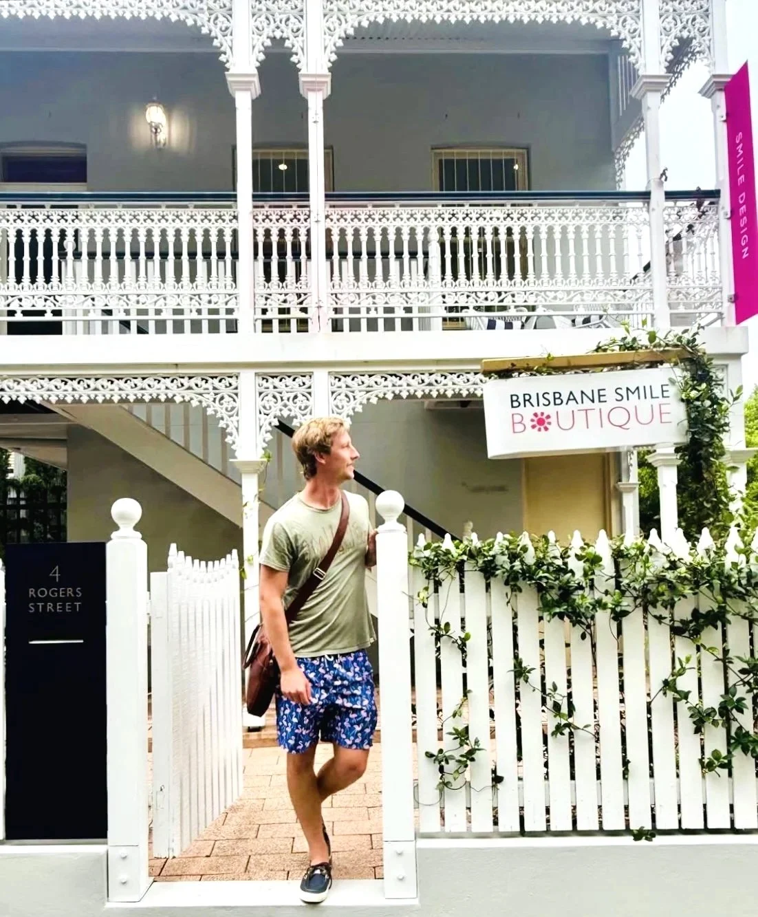 A man at Brisbane Smile Boutique standing at a white picket fence in front of a two-story building with ornate white balconies. A sign reads 'Brisbane Smile Boutique' and there is a smaller sign with the address '4 Rogers Street' in Spring Hill