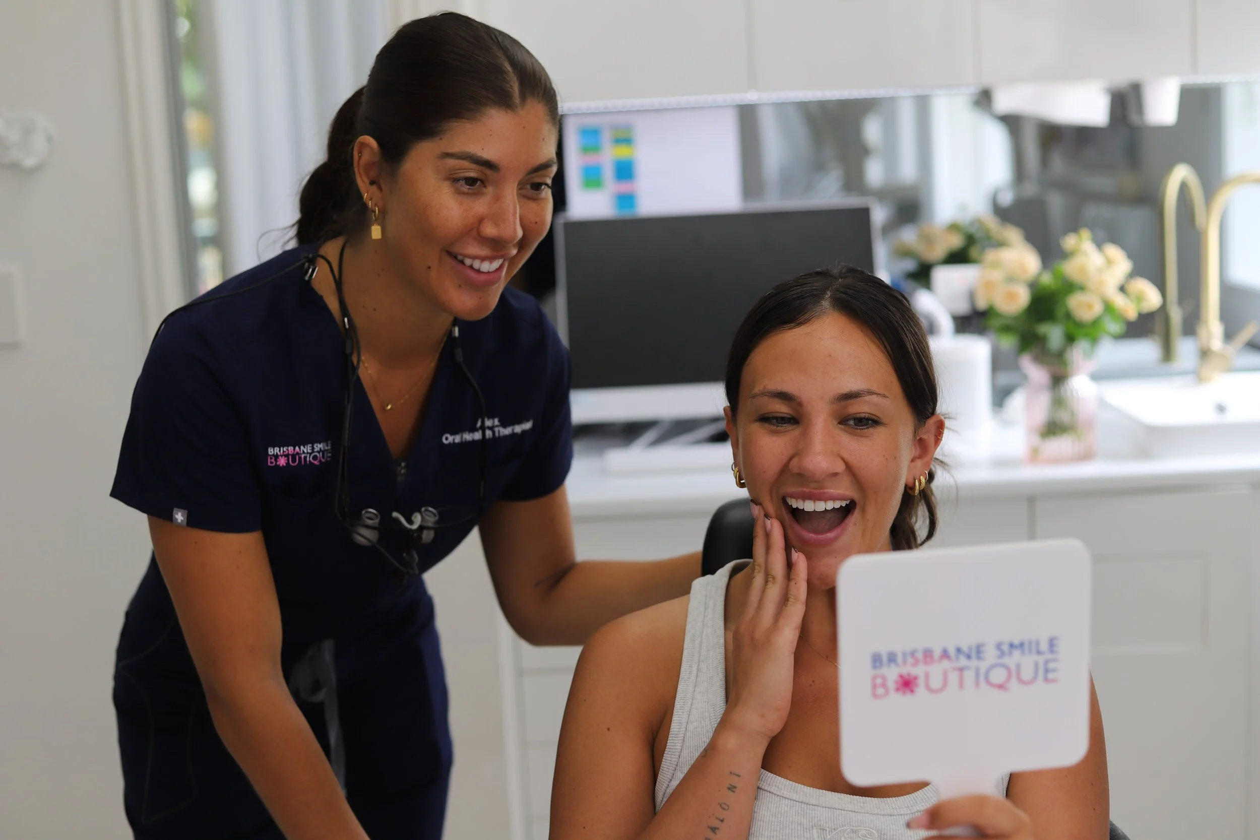 Female dental professional at Brisbane Smile Boutique, with the dentist looking into a mirror with a female patient smiling with the words brisbane smile boutique on the mirror