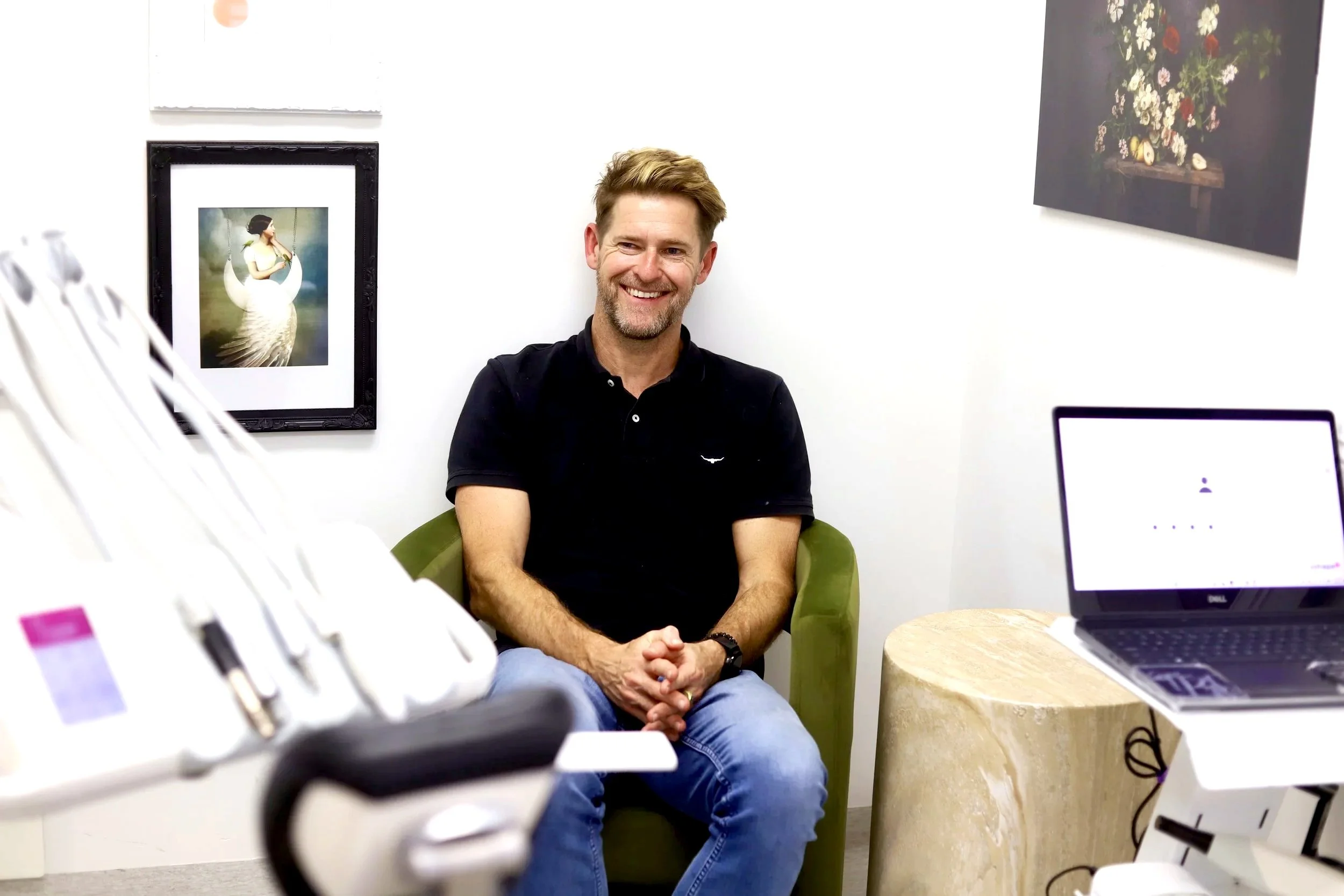 man in a dental surgery at Brisbane Smile Boutique with short hair and beard and black shirt, with computer next to him and frames behind him.
