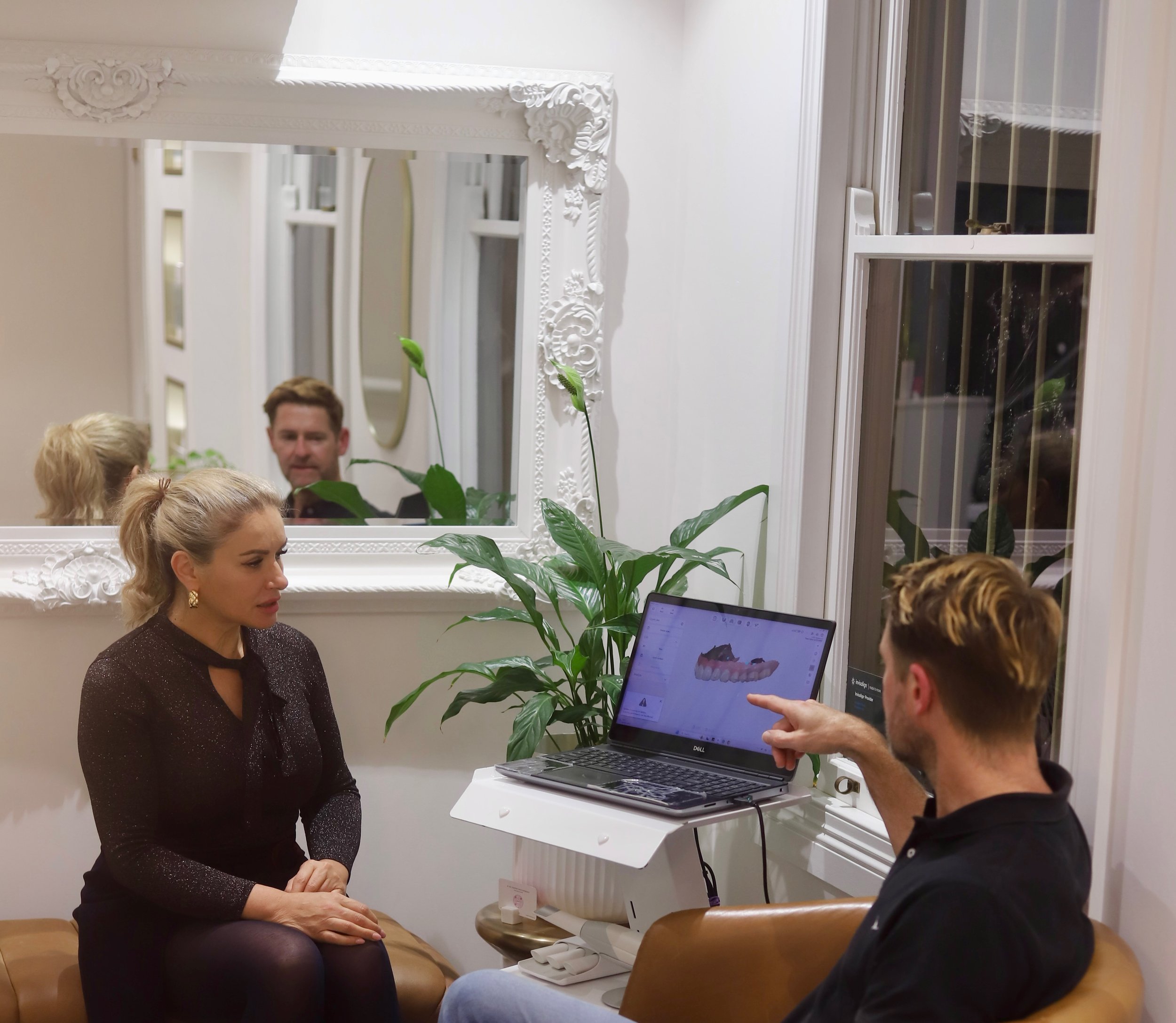 A woman sits on a brown bench during a consultation with a man pointing at a computer screen displaying a dental model. A large decorative mirror is on the wall behind them, reflecting a man’s face and the room's ceiling. A green plant is between them, and a window with bars is on the right.