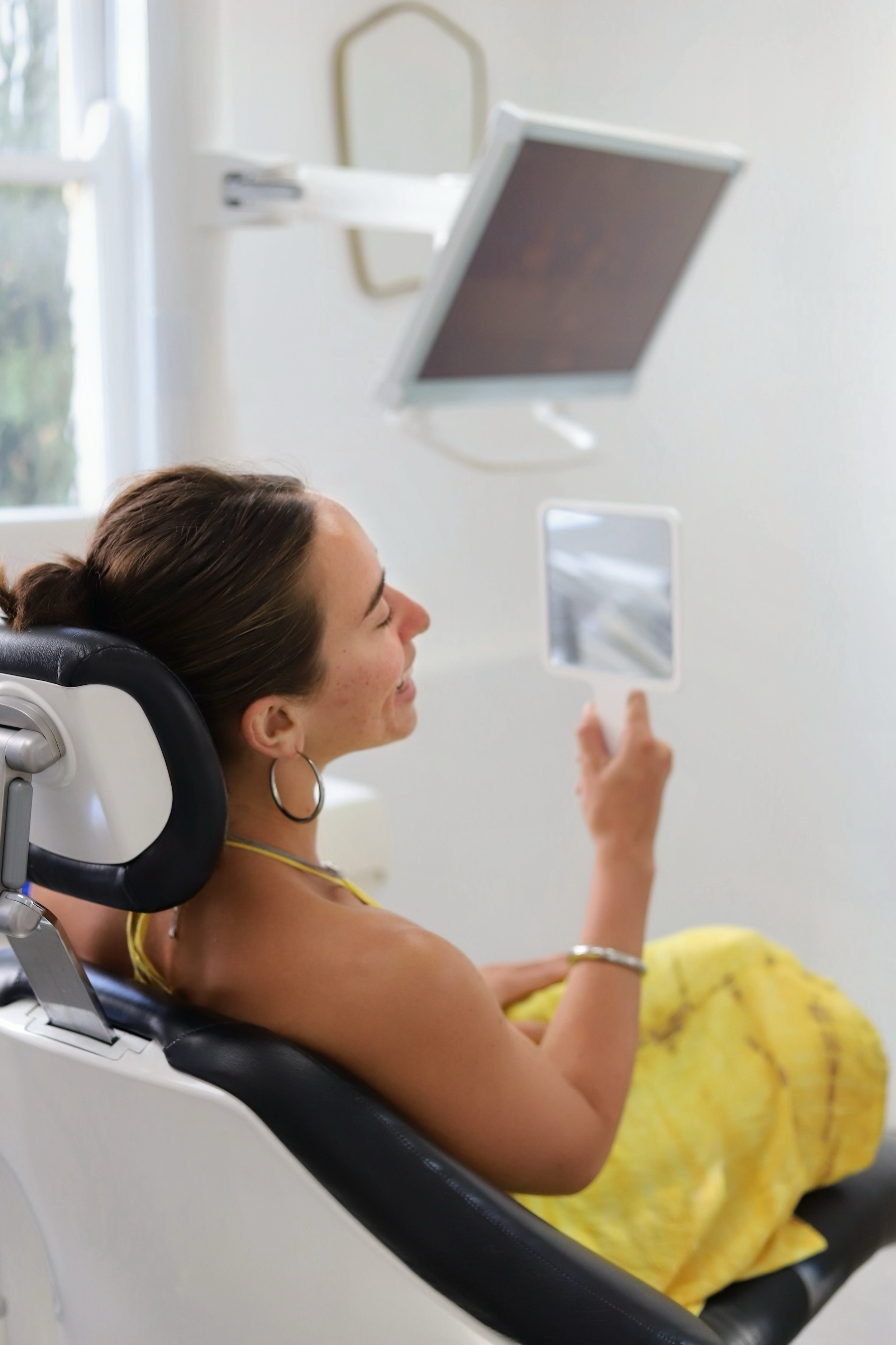 A woman at  sitting in a dental chair holding a mirror and looking at her teeth.