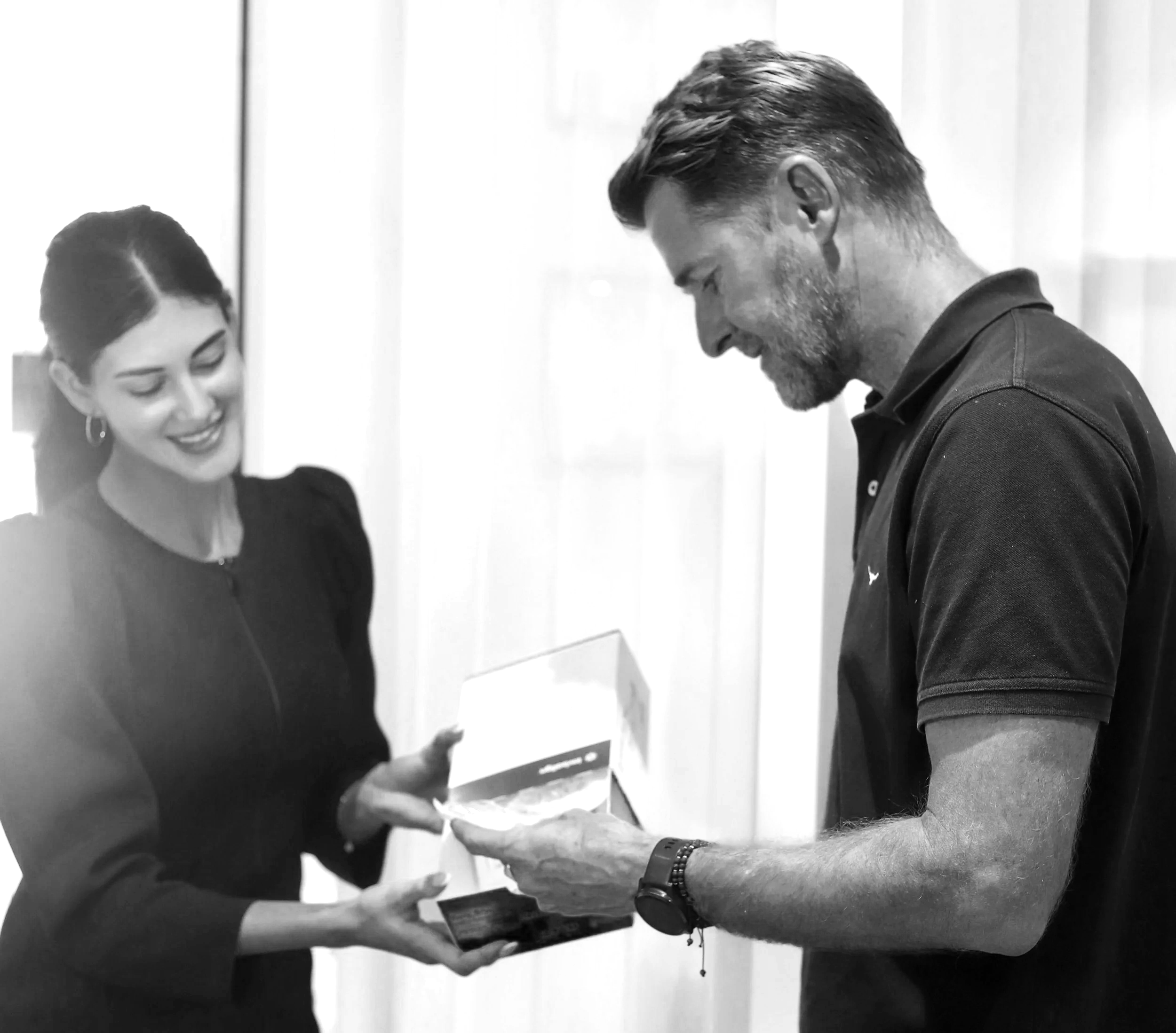 A woman is handing a male patient an Invisalign treatment box at Brisbane Smile Boutique Dental Clinic in Spring Hill, and the man is smiling and looking inside the box. They appear to be in a bright indoor setting.