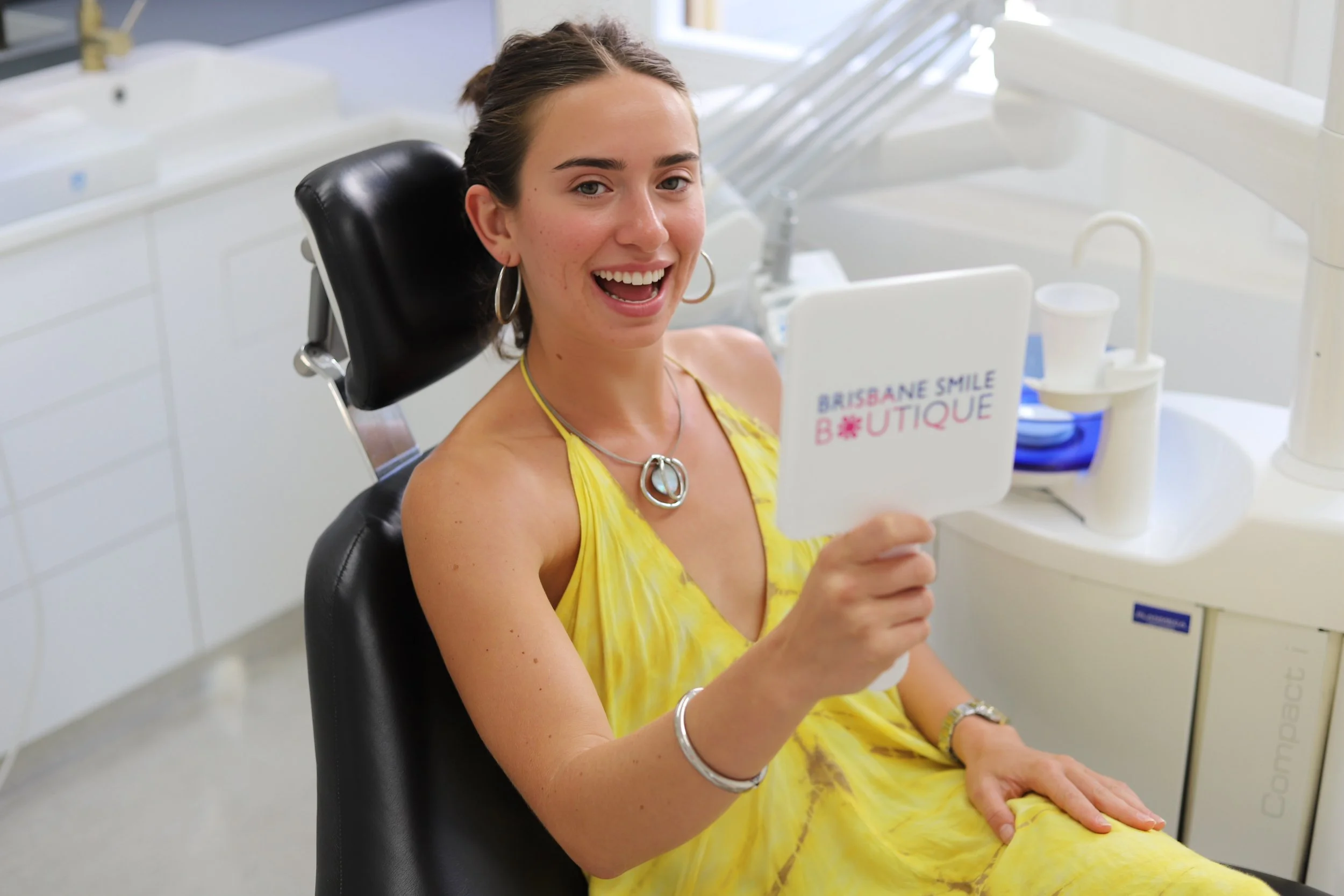 A young female in a yellow dress with a necklace on and hoop earrings smiling at the camera after cosmetic dentistry treatment at Brisbane Smile Boutique Dentistry Clinic in Spring Hill, with Dr Leila Haywood