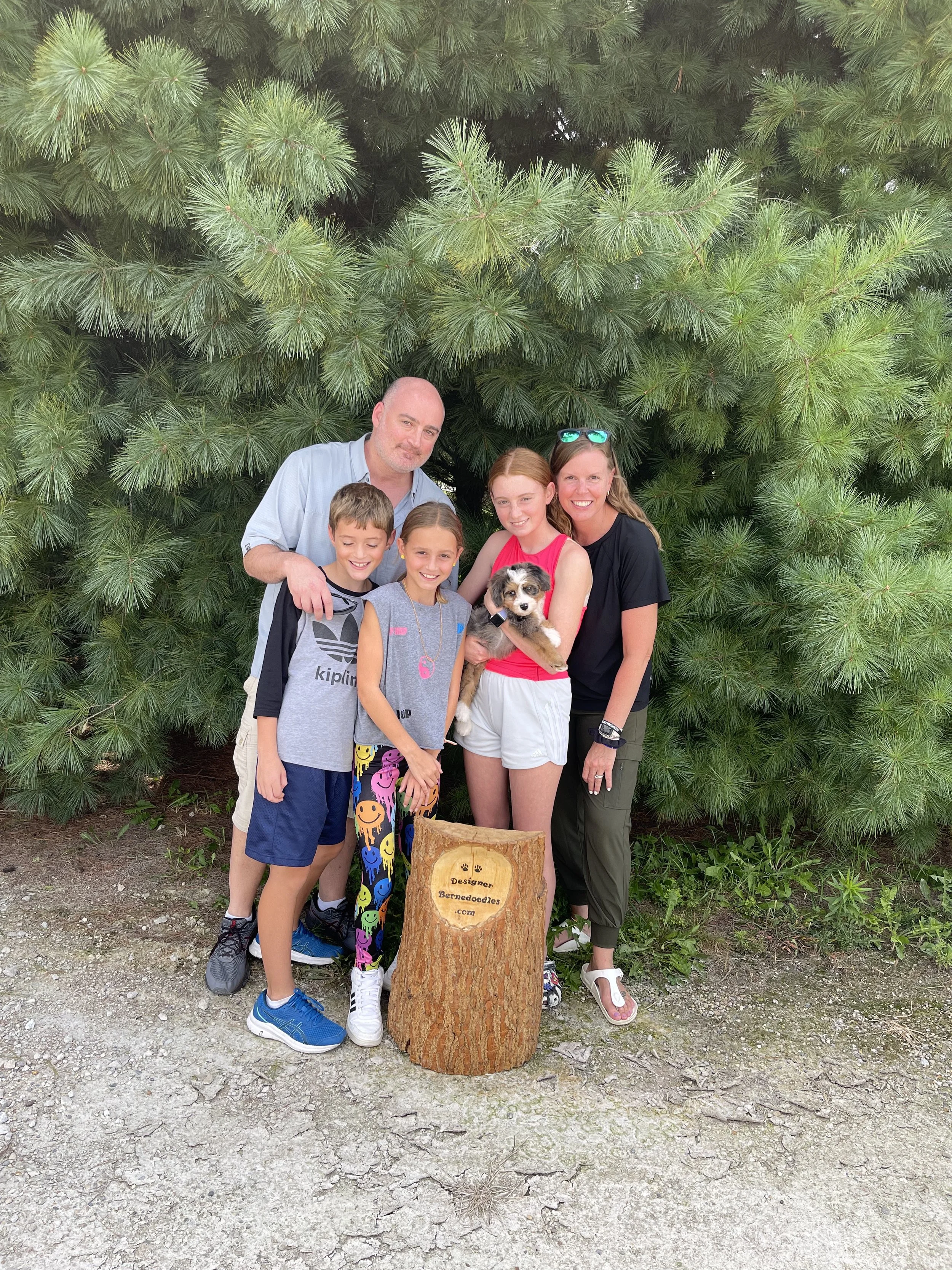 A family of five holds a hypoallergenic bernedoodle puppy outside in fornt of pine trees