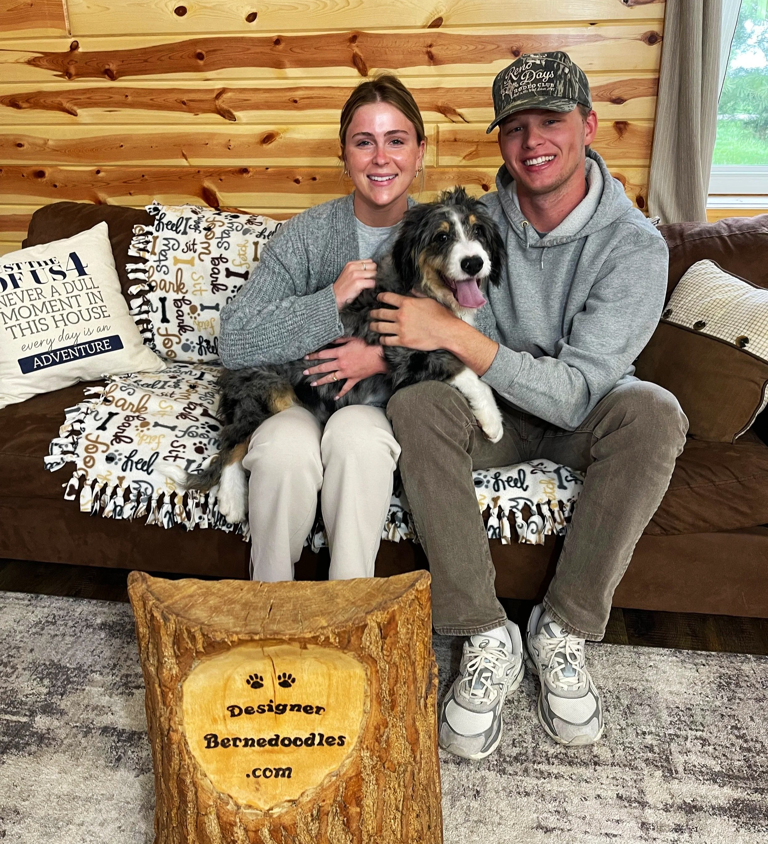 A woman and man sit on a couch holding a brown and white f1b bernedoodle