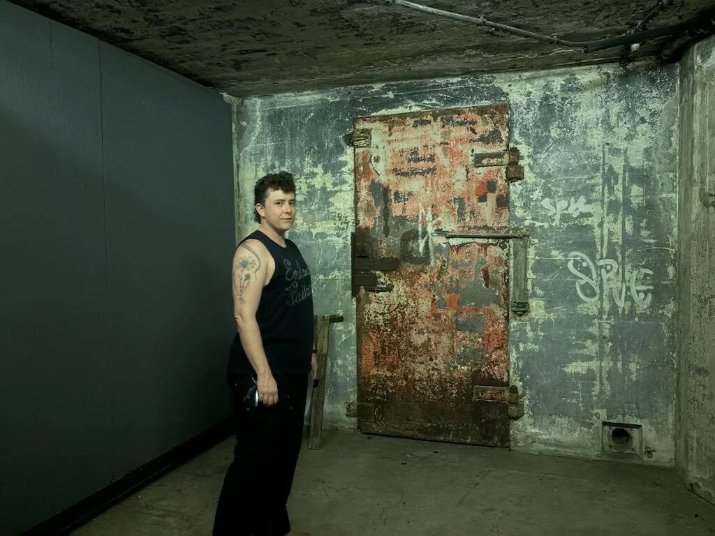Teddy Pozo looking at the camera in wide shot in 3/4 profile in an underground room. They are a white nonbinary person with curly brown hair in a short mullet. They are cleanshaven, wearing all black, and have upper arm tattoos.