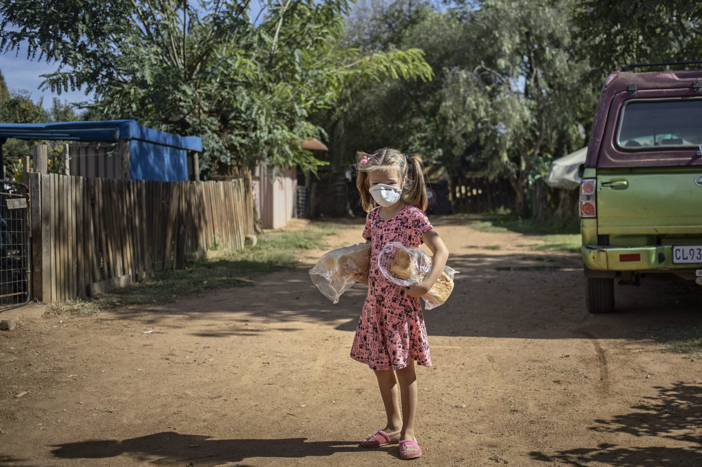 A color photograph taken during the pandemic of a young Afrikaans girl in a mask returning to her family with two loaves of bread by South African photojournalist Alon Skuy, for Conde Cotnemporary.