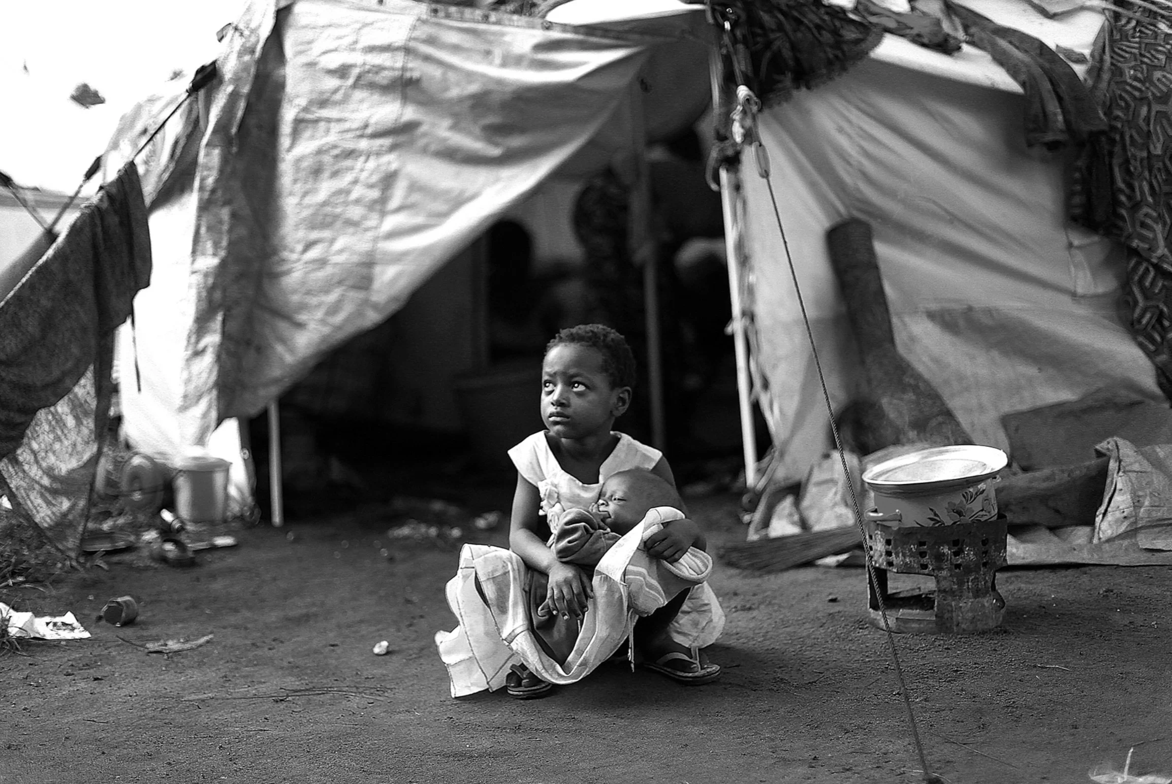 A black and white photo of a young child holding a baby outside a tent by South African photojournalist Alon Skuy, for Conde Contemporary.