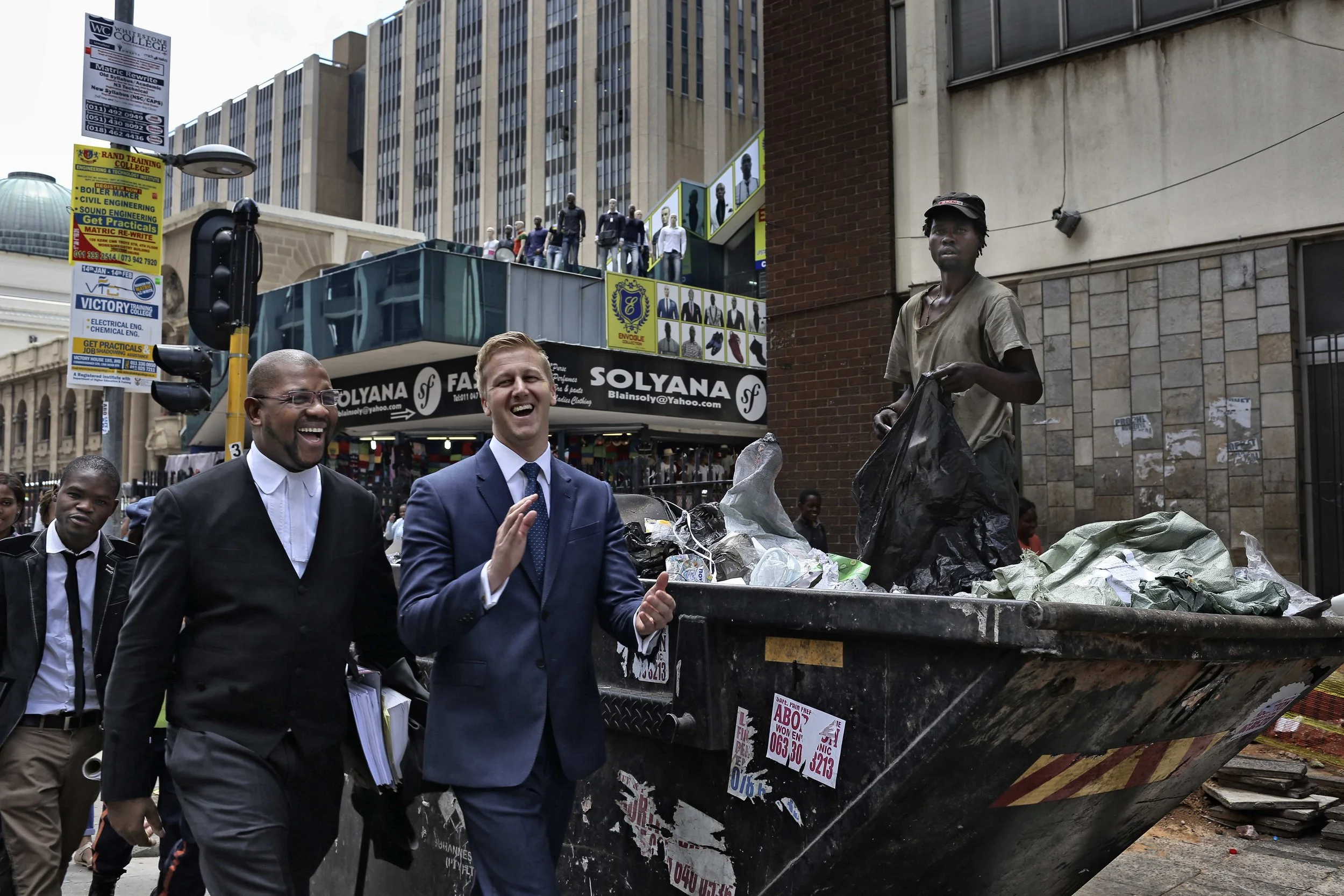 A photograph of a South African DJ and his human rights attorney laughing while walking past a man in a garbage skip putting trash in a bag by photographer Alon Skuy for Conde Contemporary.