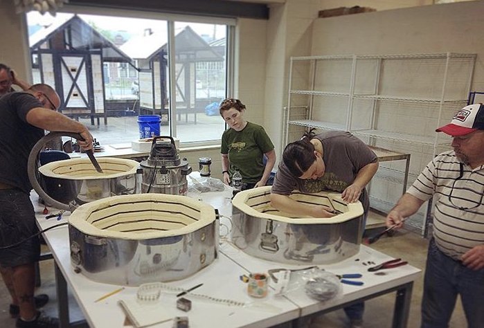 People working on large, round, metallic frames in a workshop, with tools and supplies on the table, and large windows in the background.