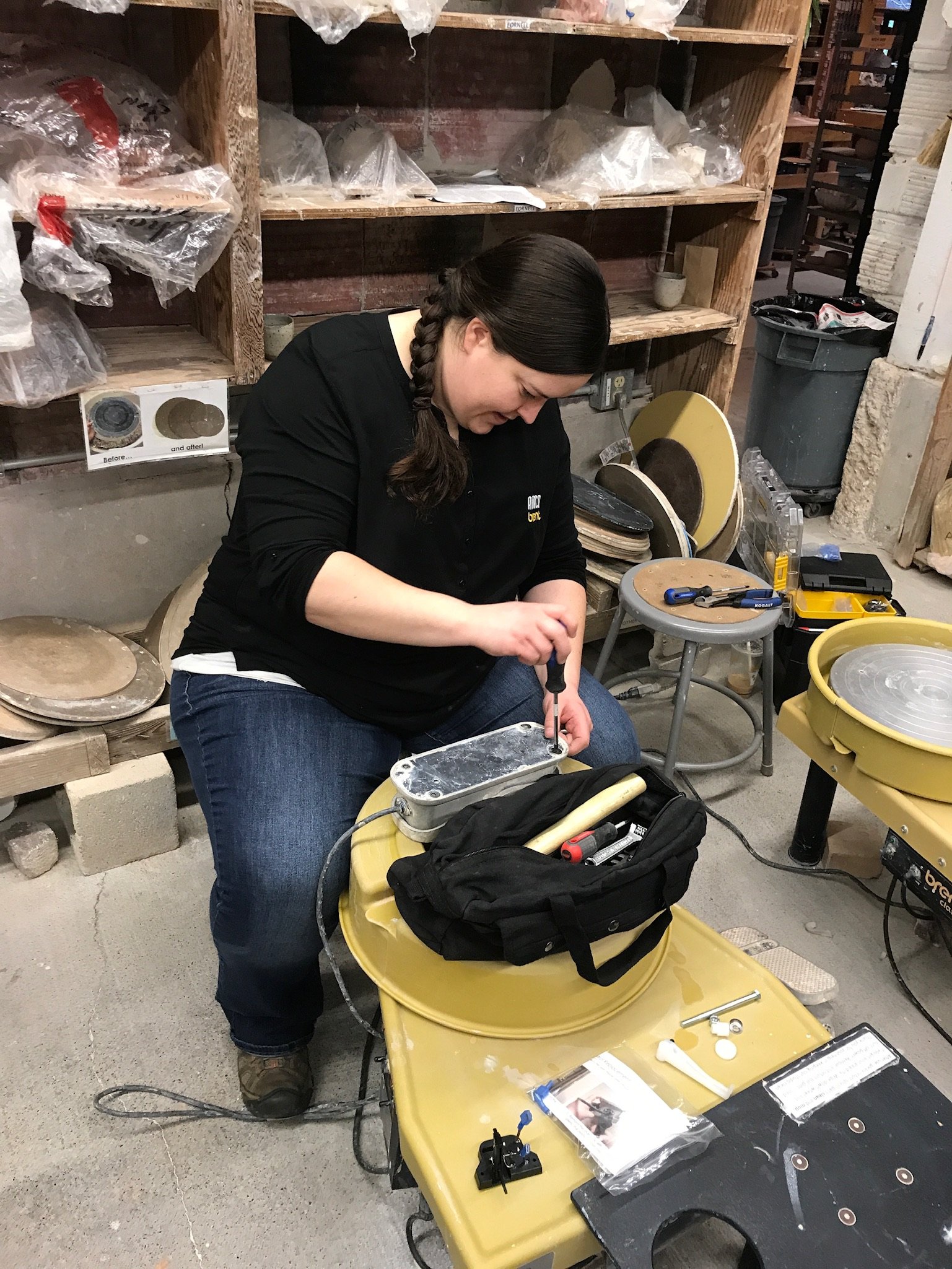 A woman working at a pottery wheel in a pottery studio, surrounded by clay and tools.