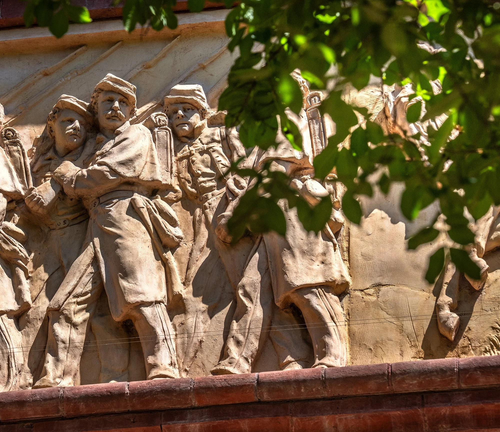 Close-up view of a stone relief sculpture depicting soldiers with military gear, partially obscured by green leaves in the foreground.