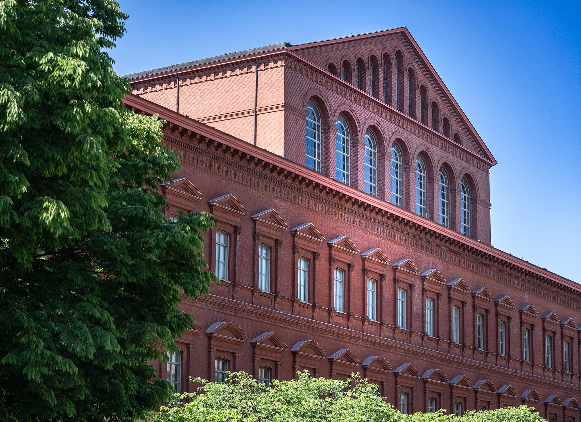 A red brick building with large arched windows, tall trees with green leaves in front, and a clear blue sky in the background.