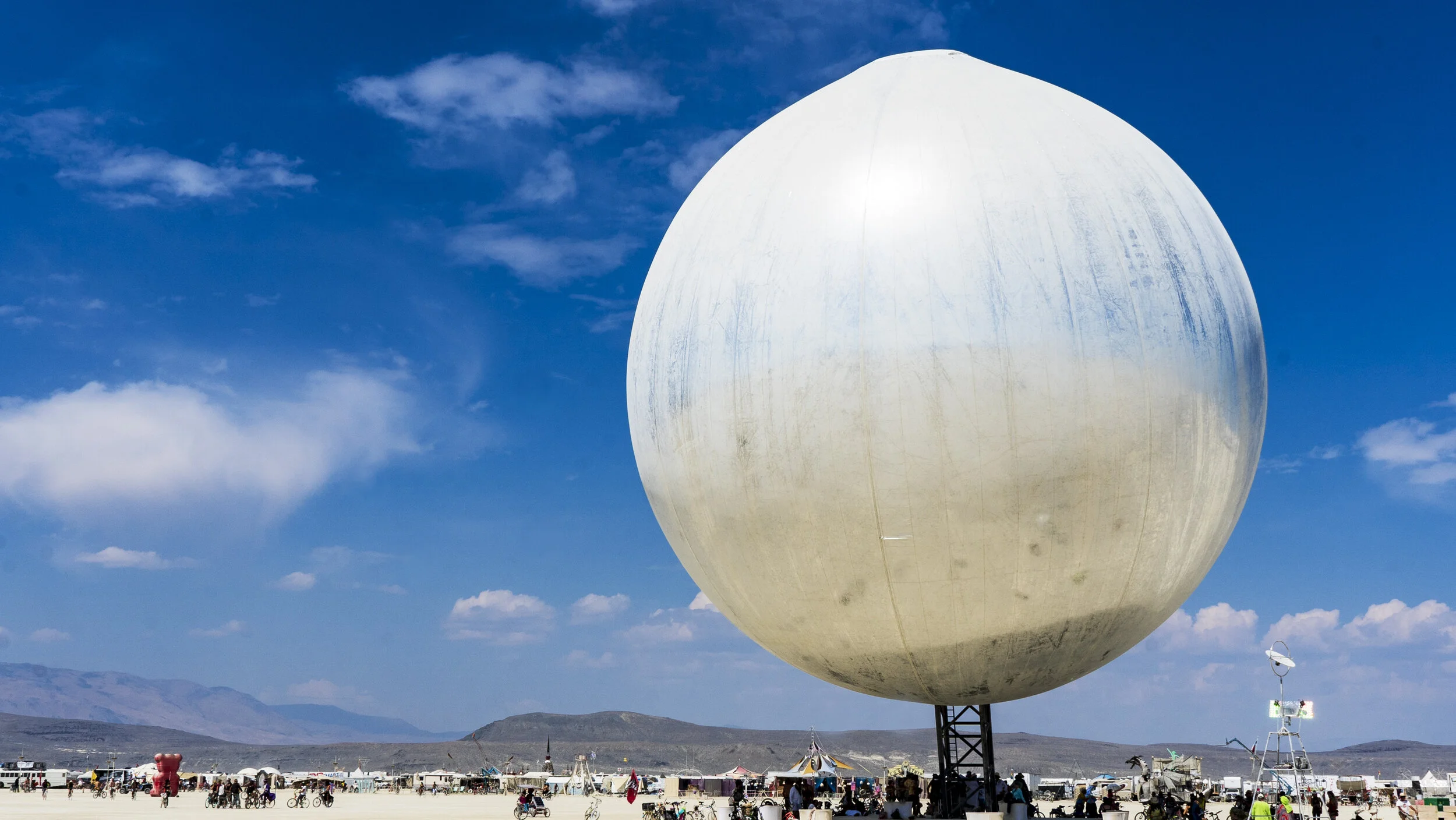 Large spherical structure at a desert event with people and bicycles, under a blue sky.