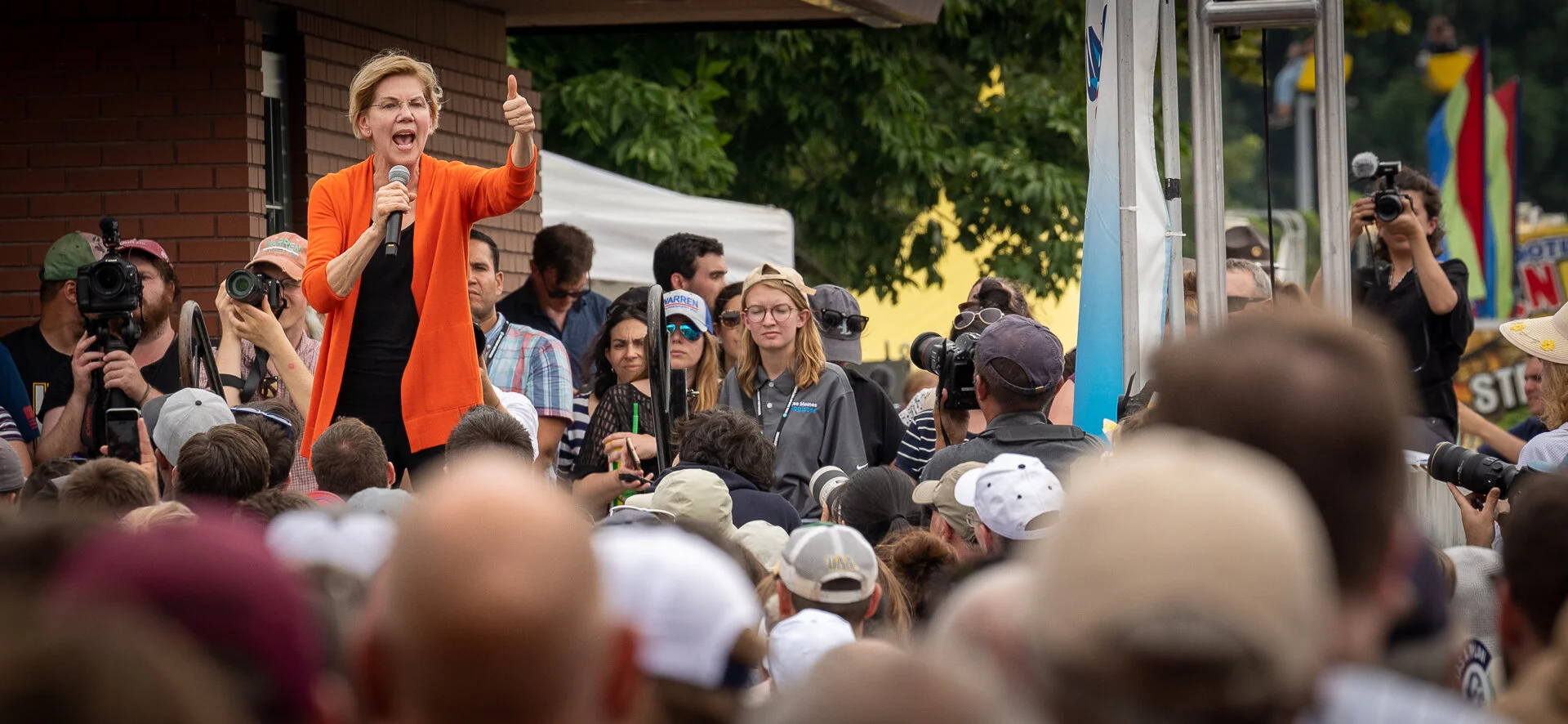 Woman in orange jacket speaking to a crowd outdoors with several people taking photos and videos.