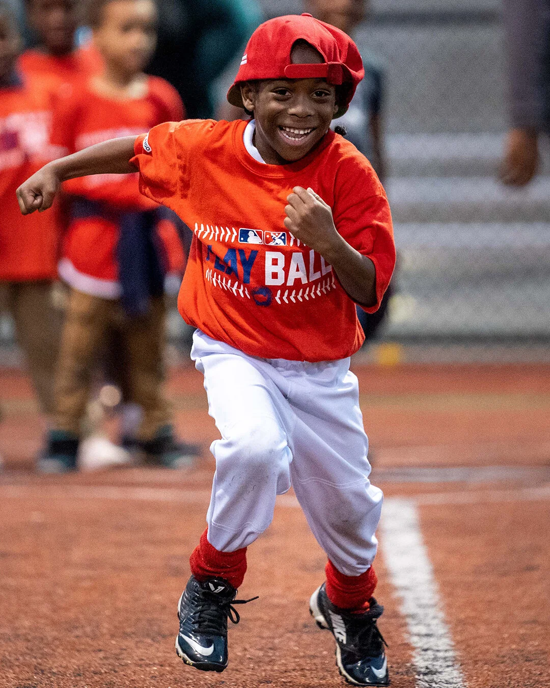 Smiling boy wearing a red baseball cap and shirt, running on a field.