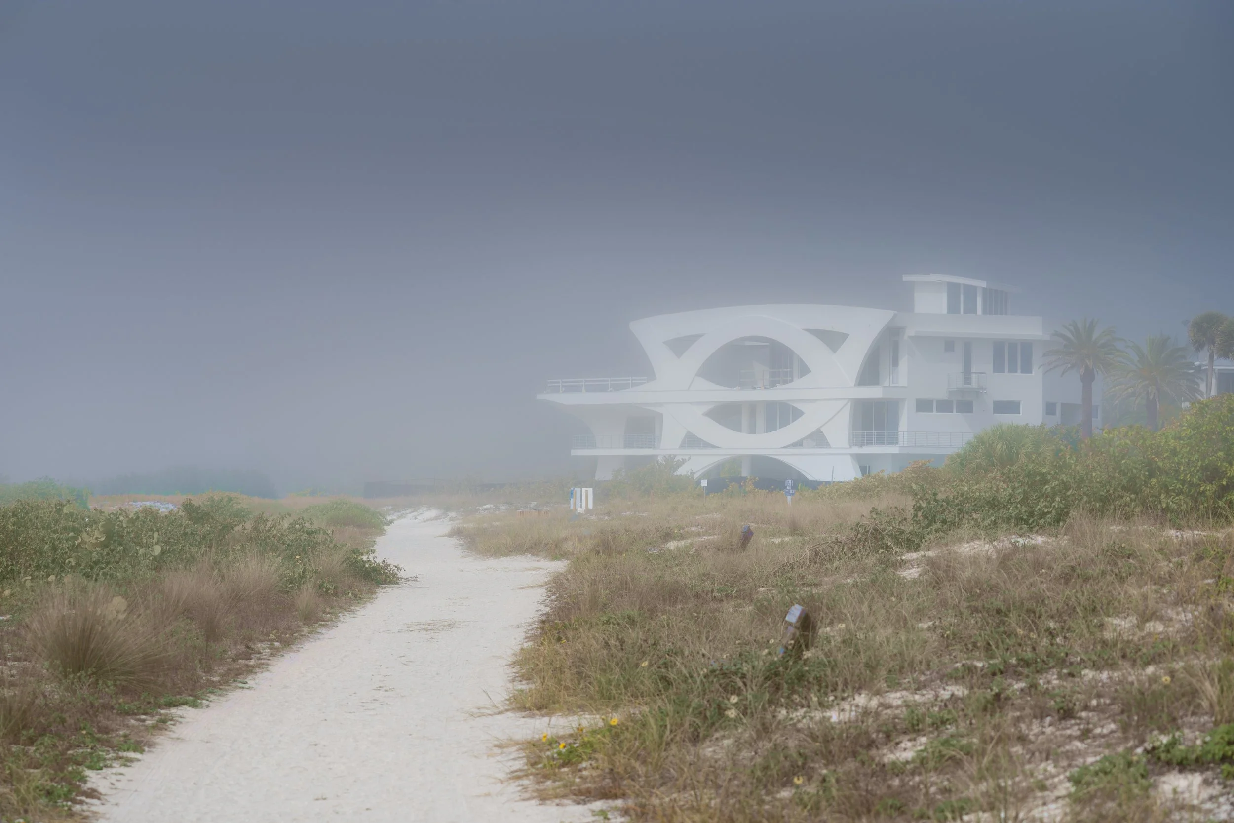 Modern white house with unique circular architecture in foggy beach setting with sandy path and vegetation.
