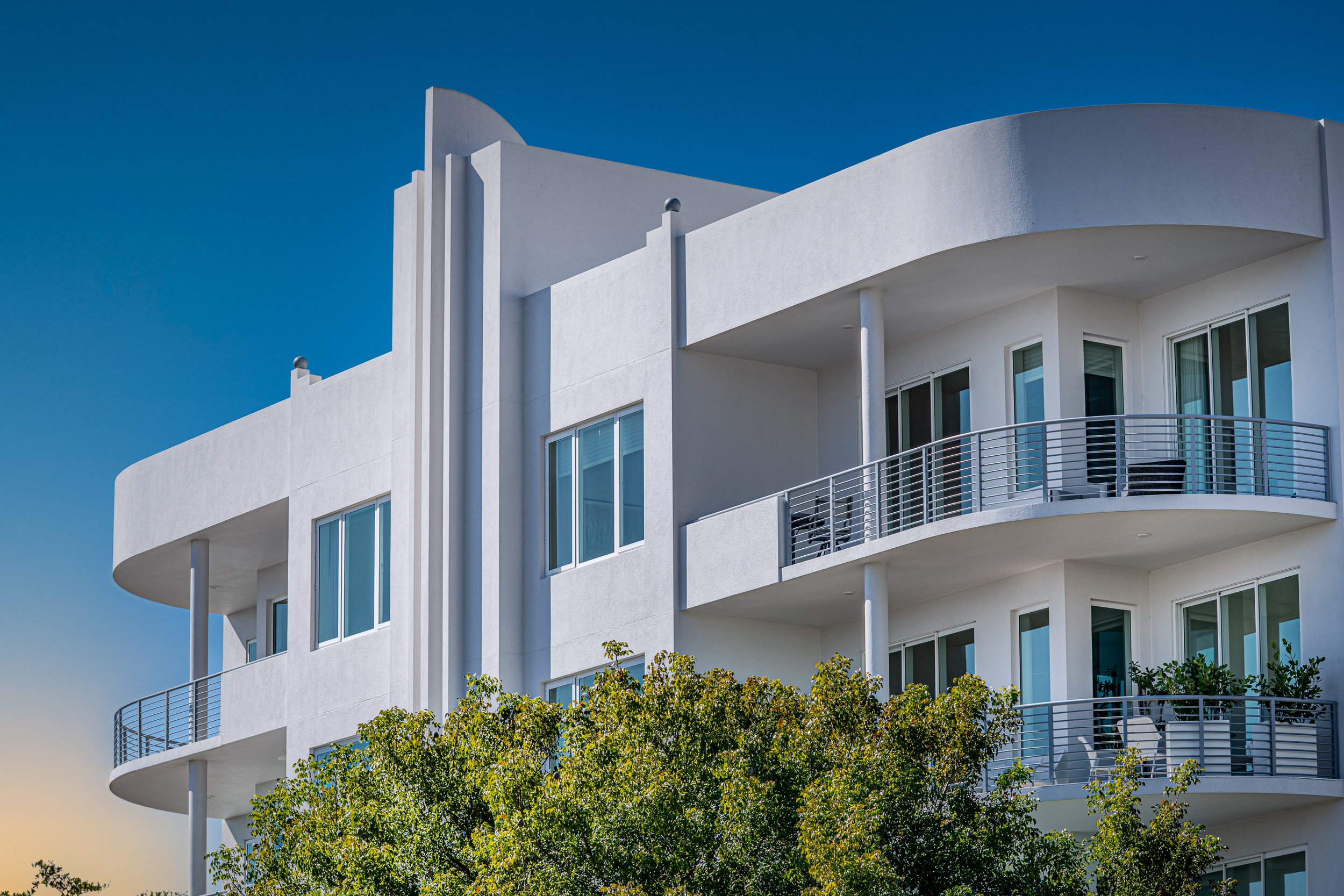 Modern white building with curved balconies and large windows, surrounded by green trees under a blue sky.