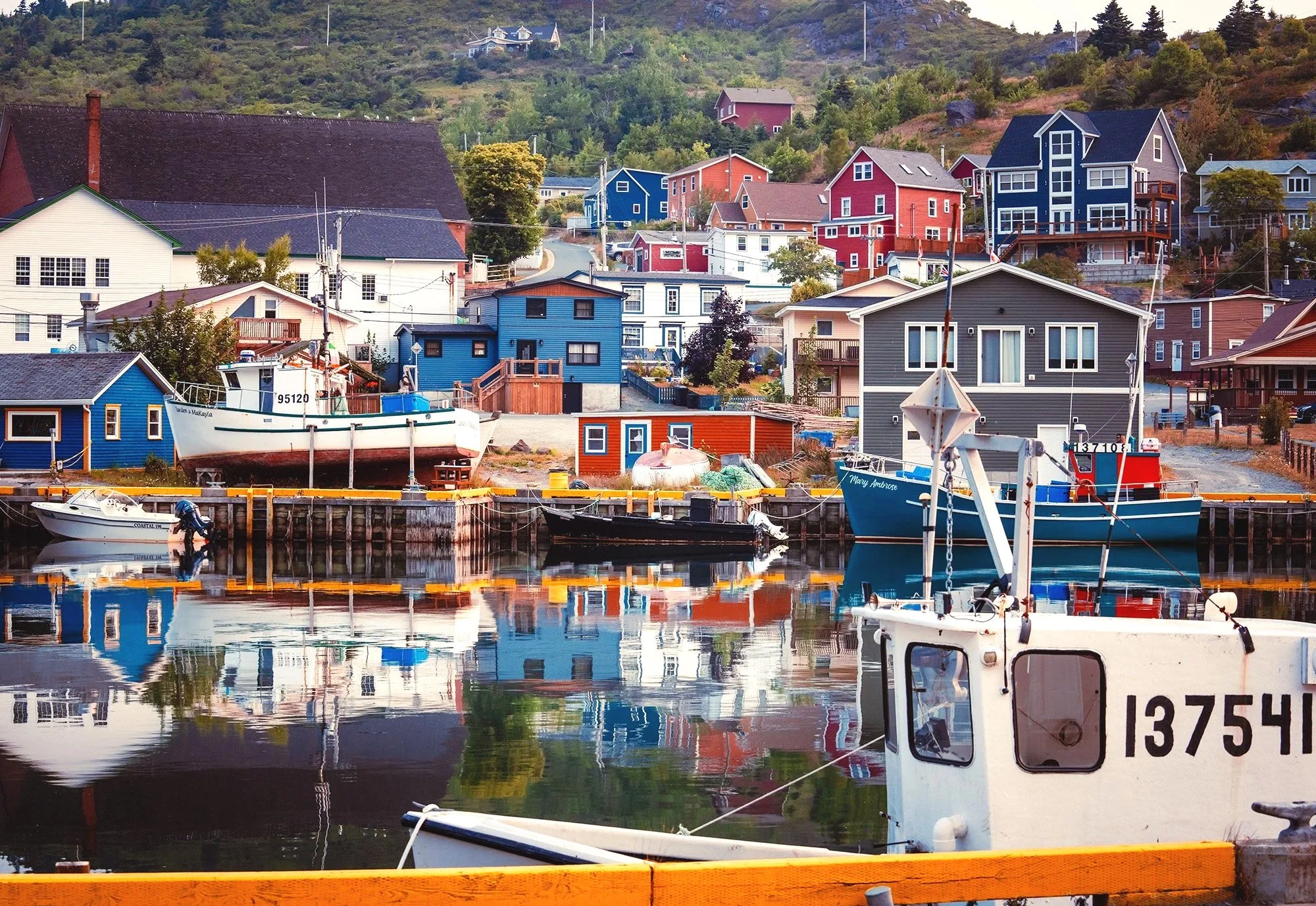 A scenic harbor with several colorful houses on a hillside and boats docked in calm water, reflecting the buildings and boats.
