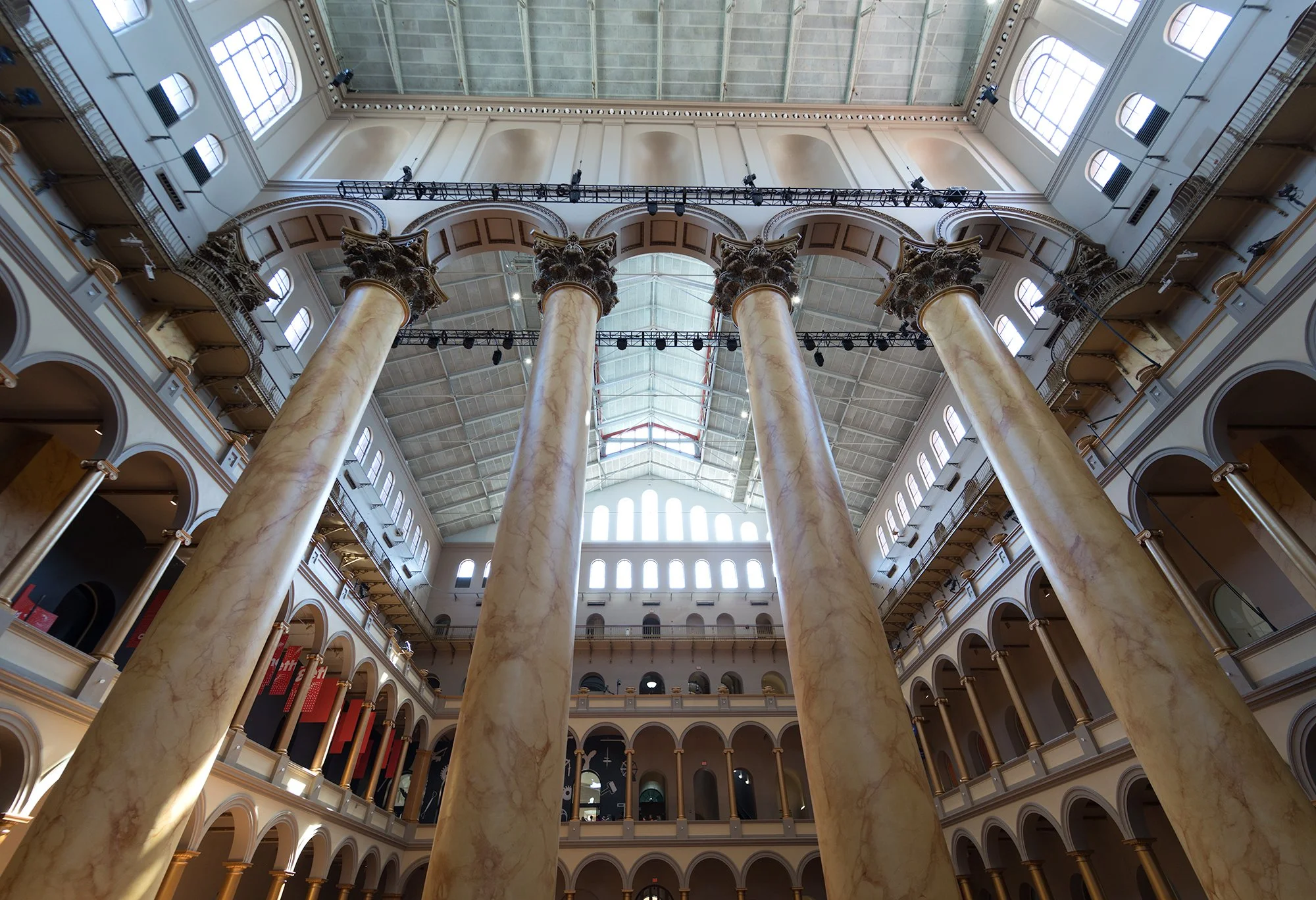Interior view of a grand building with tall marble columns, multiple floors with archways, and a high ceiling with large windows allowing natural light.