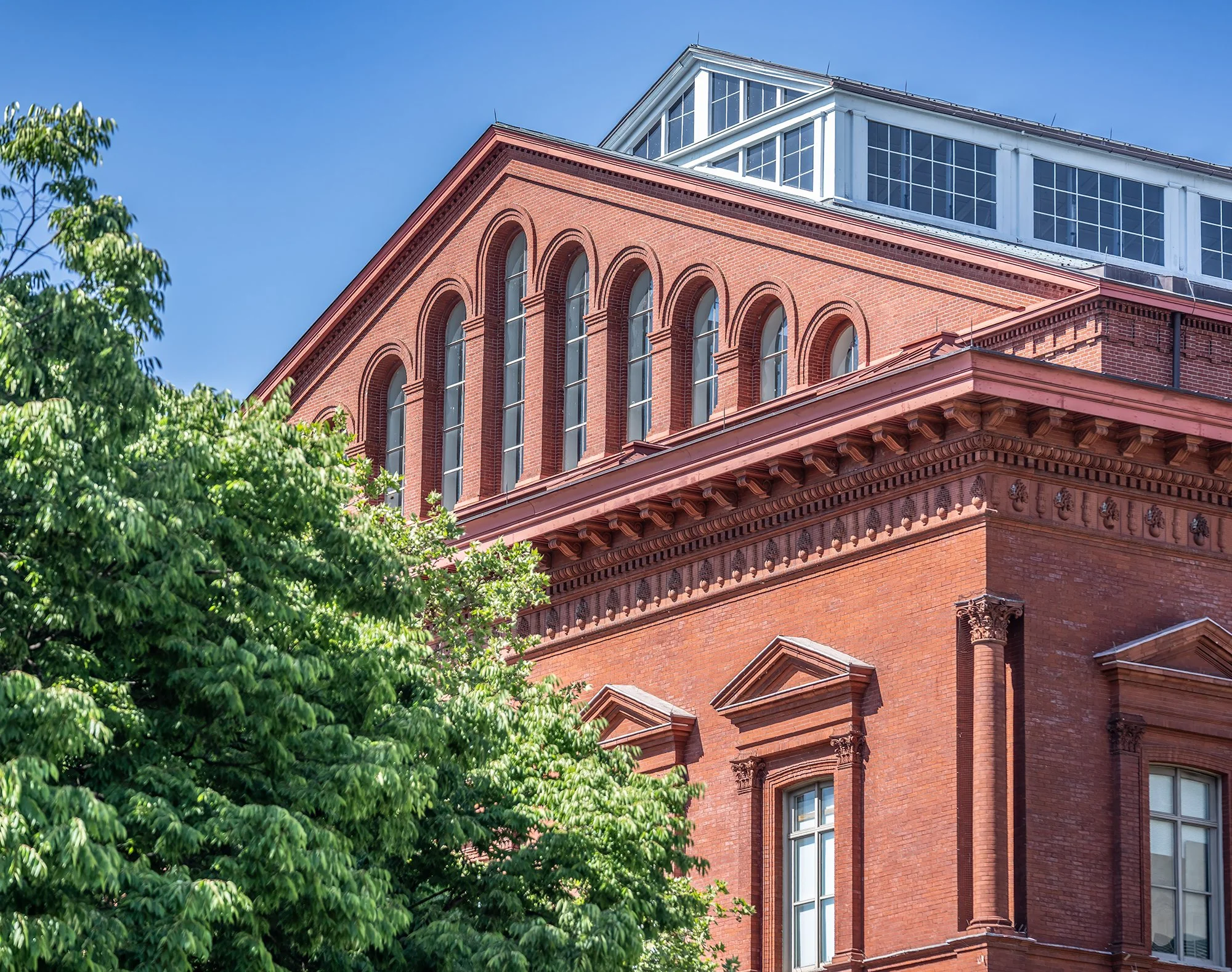 The image shows the upper part of a historic brick building with classical architectural details, large arched windows, and a glass enclosed rooftop area. In the foreground, there are lush green trees, and the sky in the background is clear and blue.