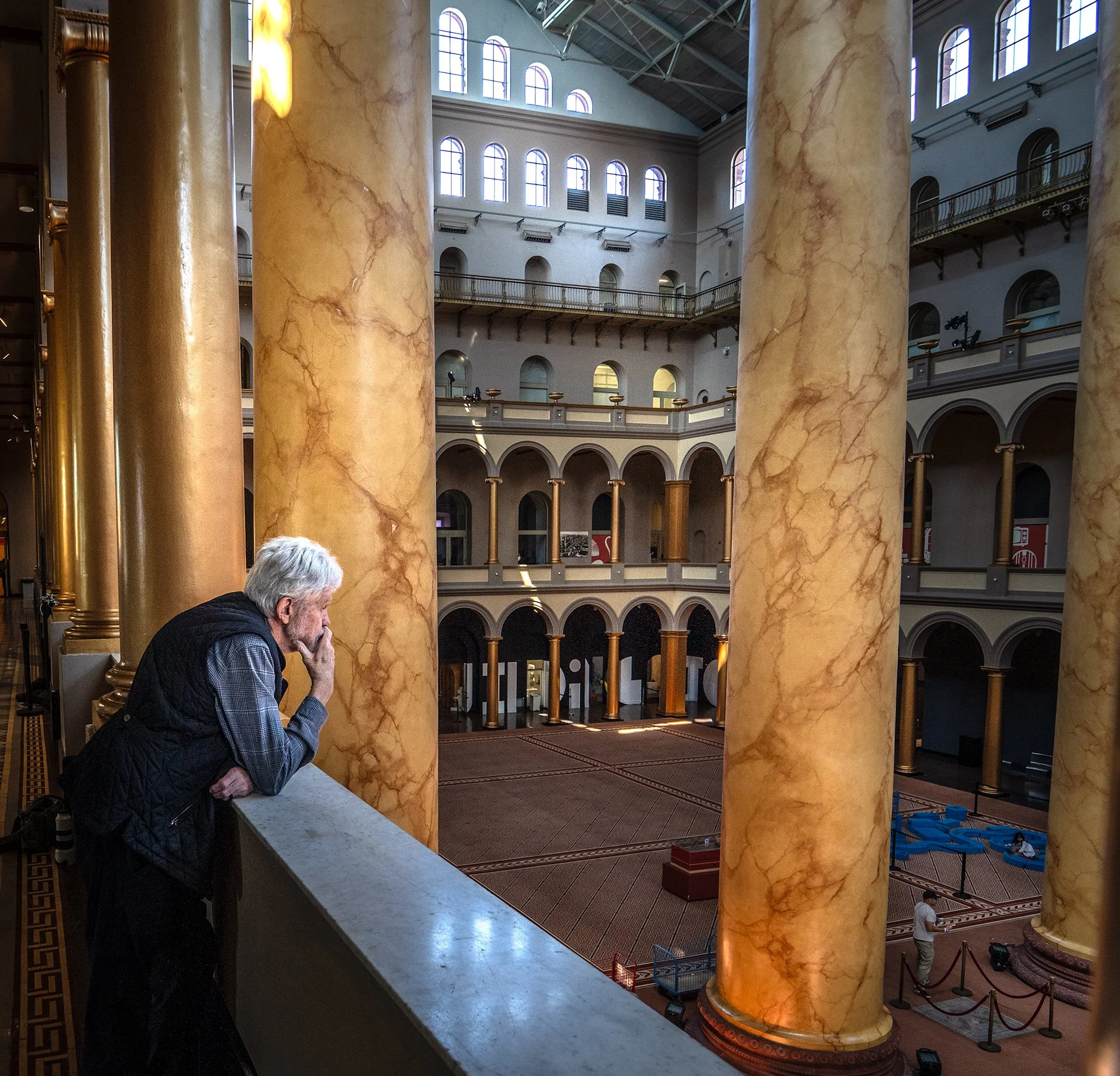 Interior of a grand building with large marble columns, multiple floors with arches and railings, and natural light coming through numerous upper windows. A man with gray hair, glasses, and a black vest is leaning on a ledge, looking downward.