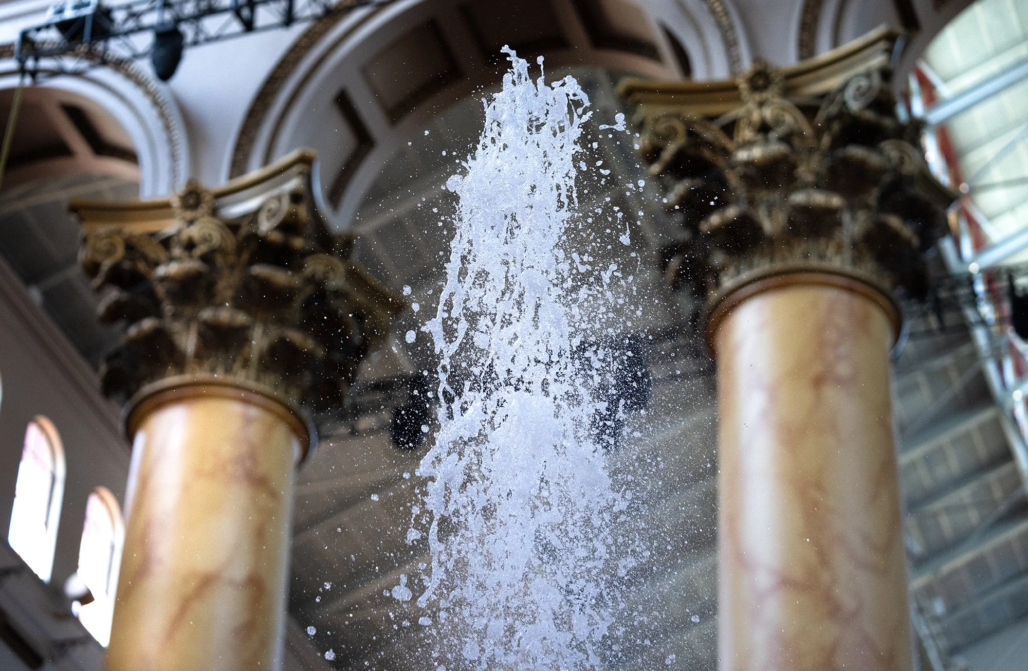 Water fountain with water splashing upward, located indoors with decorative columns and arches.