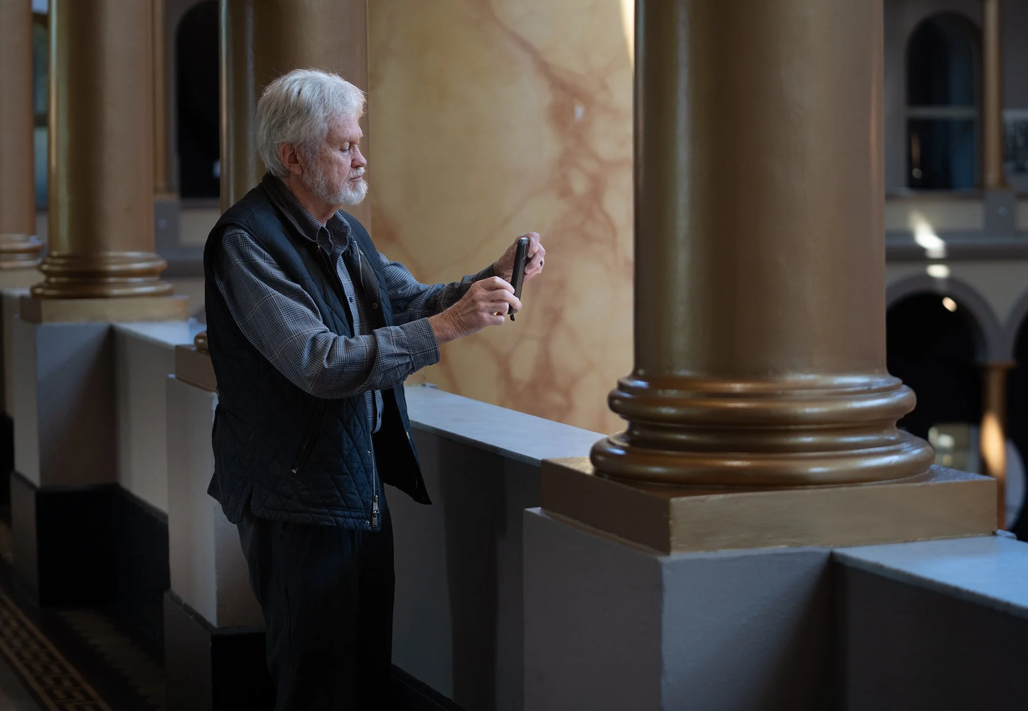 An elderly man with gray hair and beard standing at a counter in a building with large decorative columns, looking at his phone.
