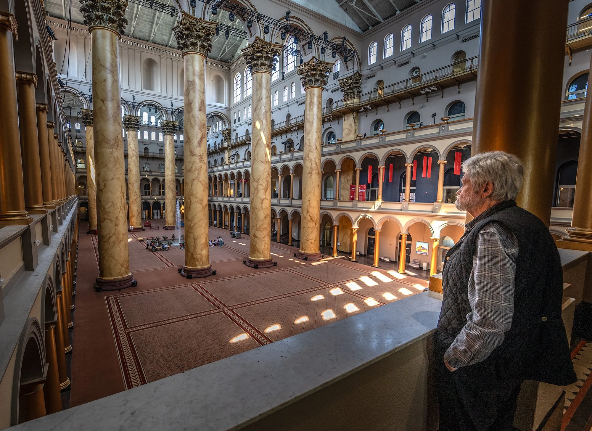 Interior view of a large, historic building with tall marble columns, arched windows, and multiple floors with balconies. A man with gray hair and a checkered shirt is standing and looking at the architecture.